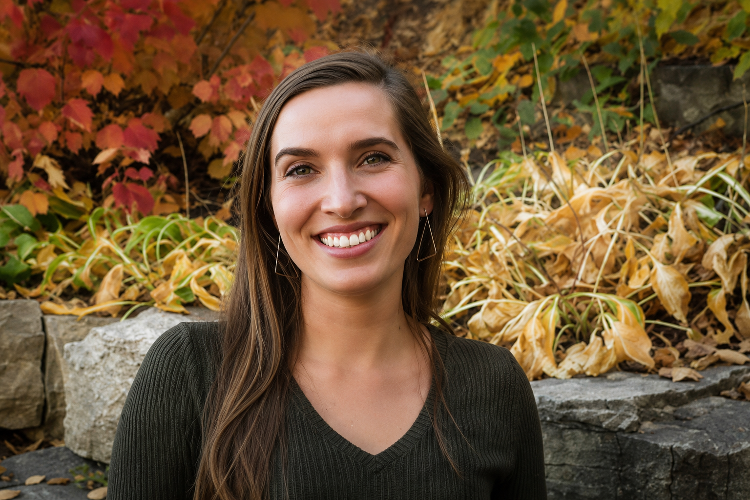 A smiling woman with long brown hair, wearing a dark v-neck sweater and geometric earrings, standing outdoors in front of autumn leaves and rocks.