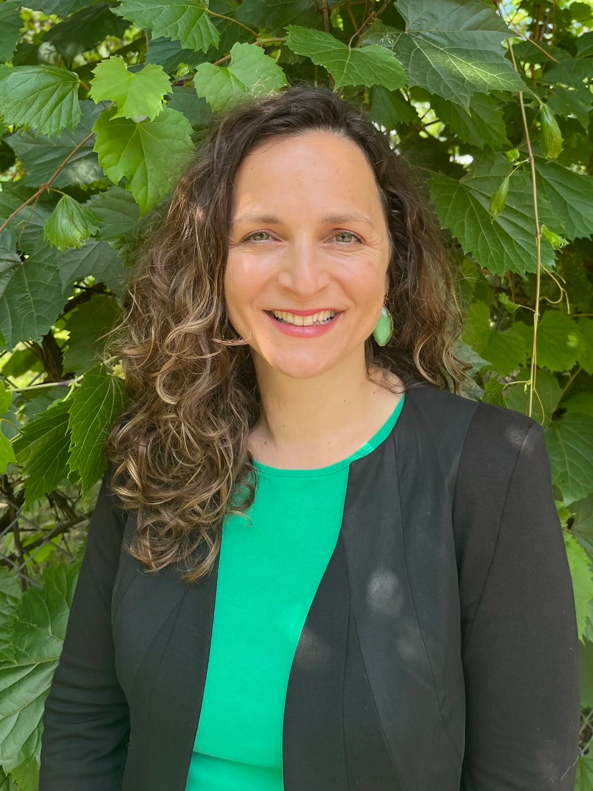 A woman with curly brown hair wearing a green top and black blazer, smiling, standing in front of green leafy background.