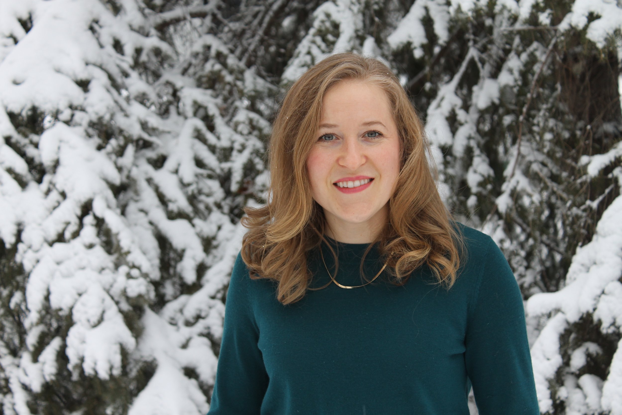 A woman with long, wavy, blonde hair and blue eyes smiling outdoors in front of snow-covered trees.