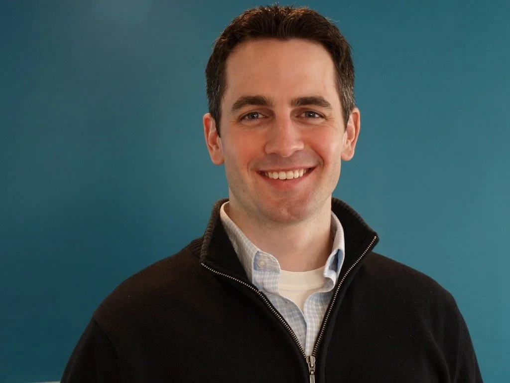 A smiling man with dark hair, wearing a white collared shirt and a black zip-up sweater, standing in front of a blue background.