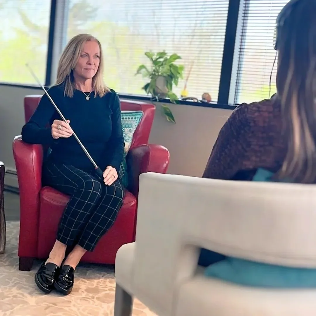 A woman with blonde hair sitting on a red armchair, holding a pointer, in a bright office with large windows and blinds, speaking to another person with long hair who is seated in a white chair.