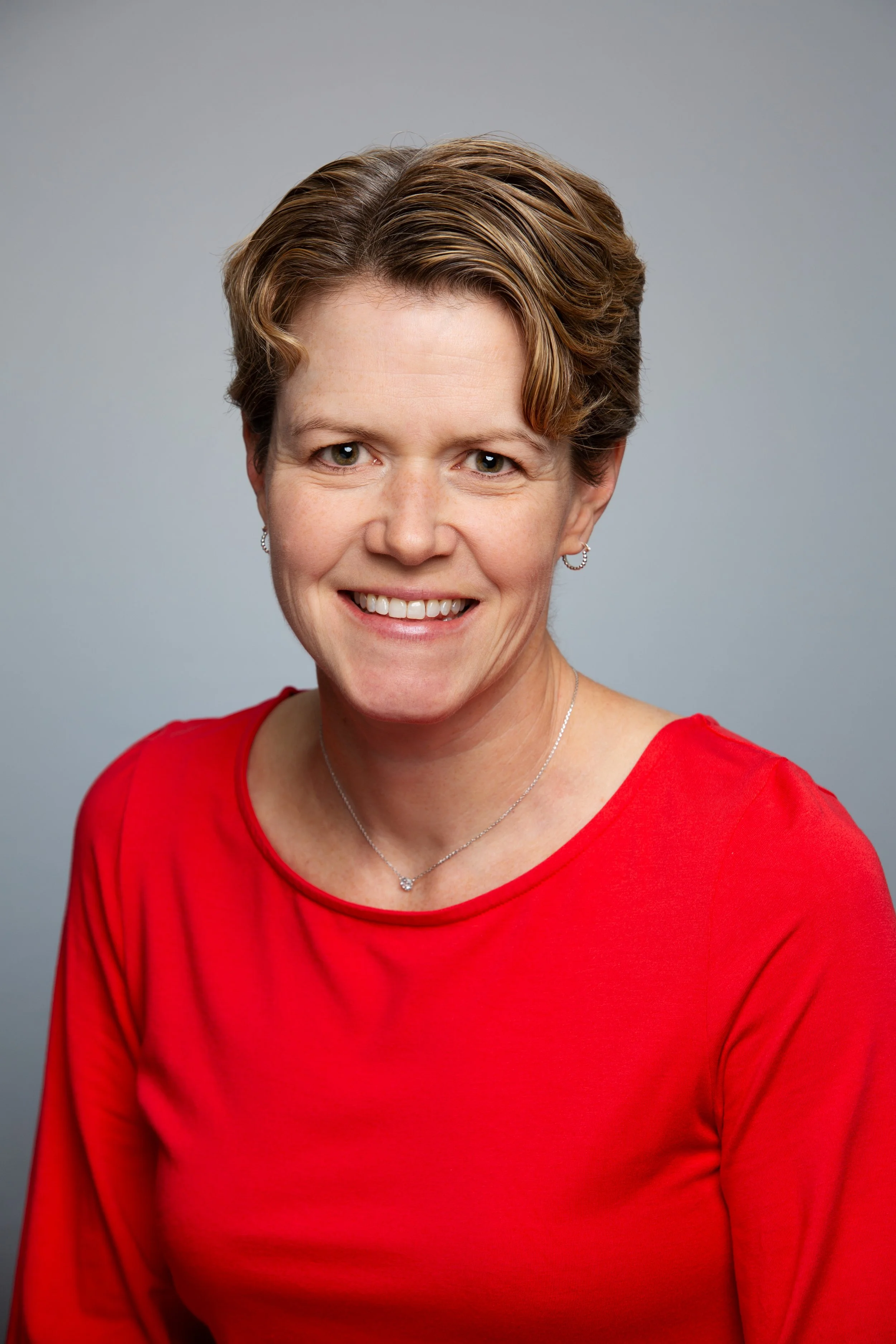 A woman with short brown hair, wearing a red top, smiling, with a light gray background.