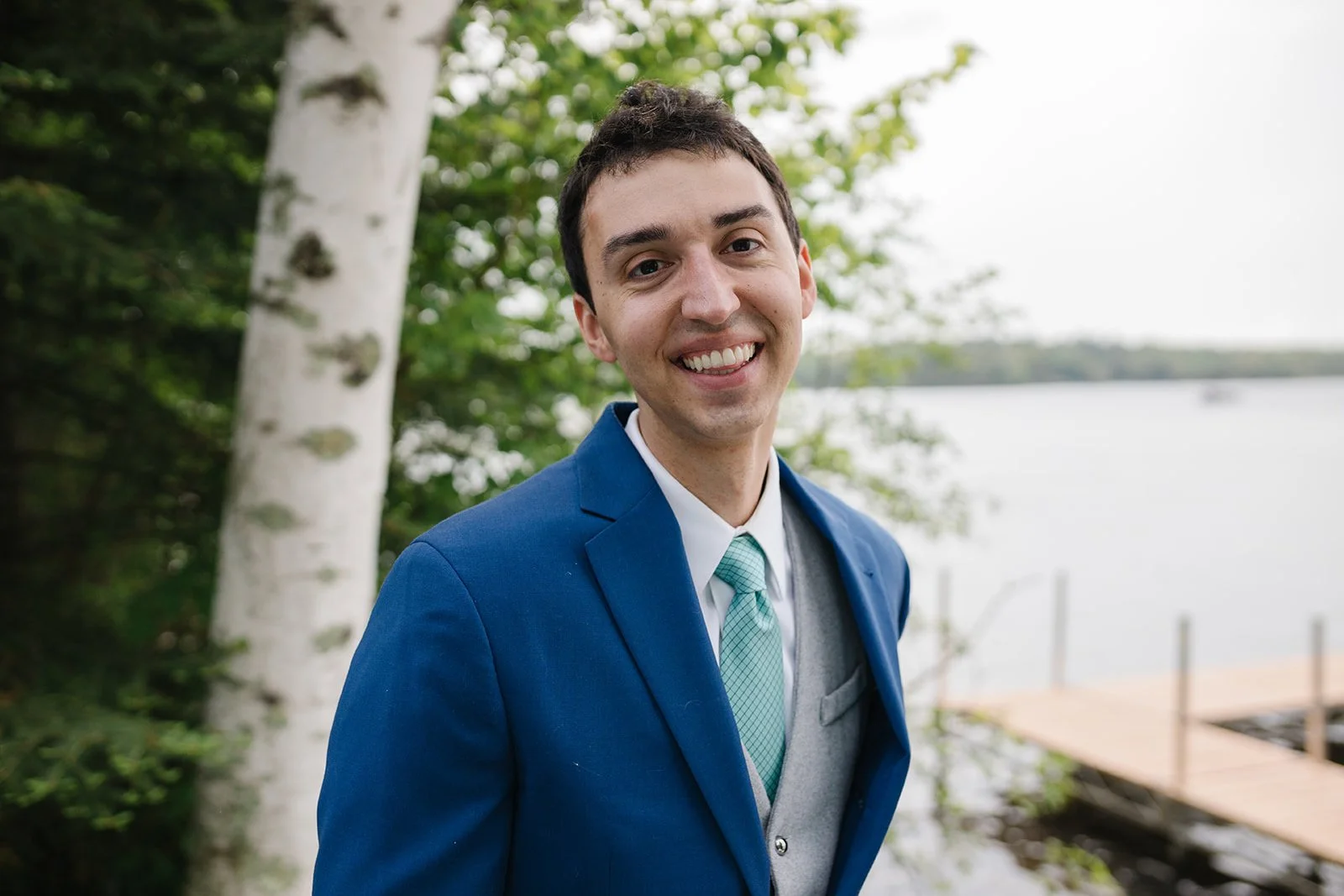 A young man smiling outdoors near a body of water with trees and a dock in the background.