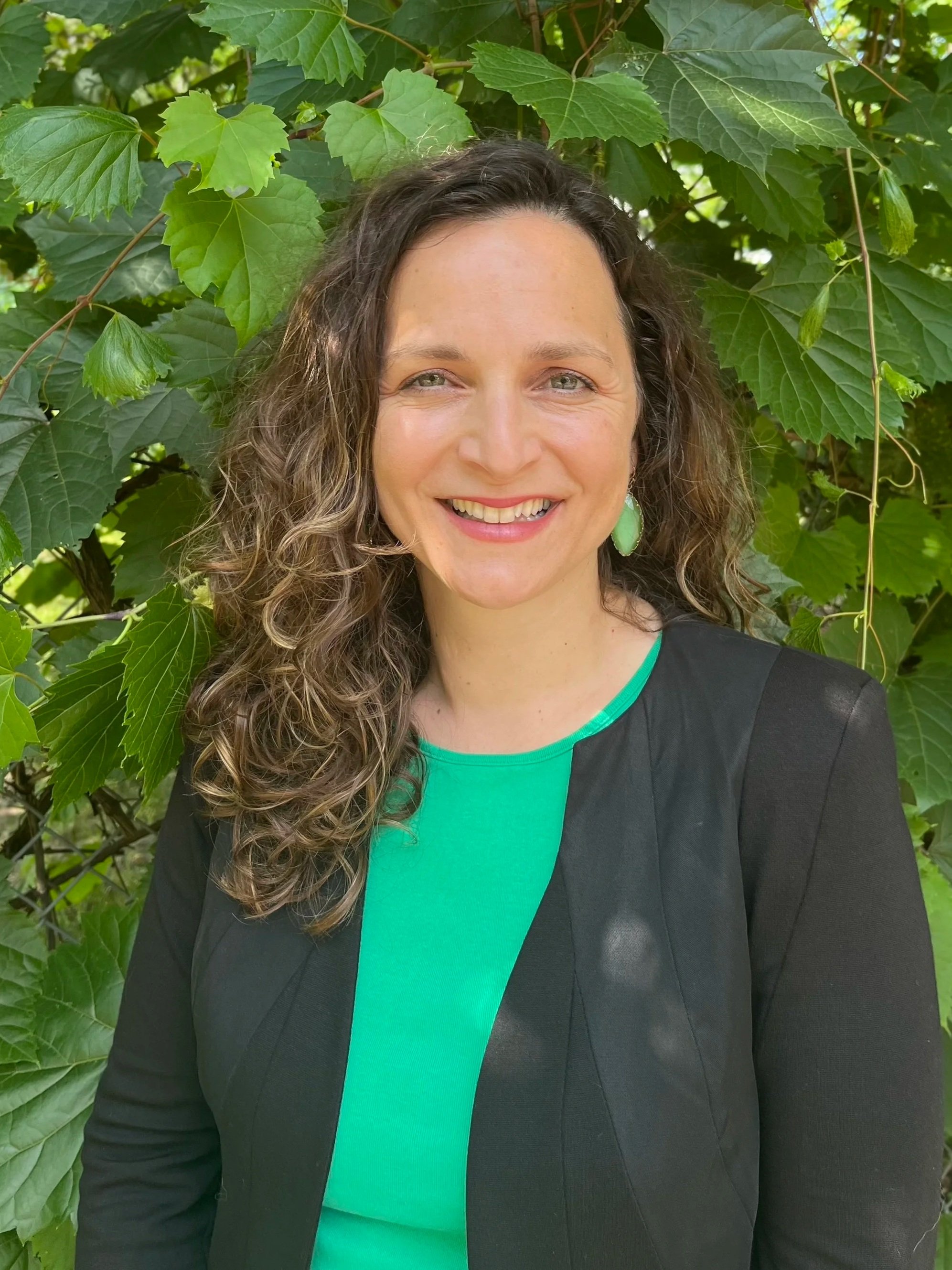 A woman with curly brown hair and green earrings standing in front of green leaves, smiling at the camera.