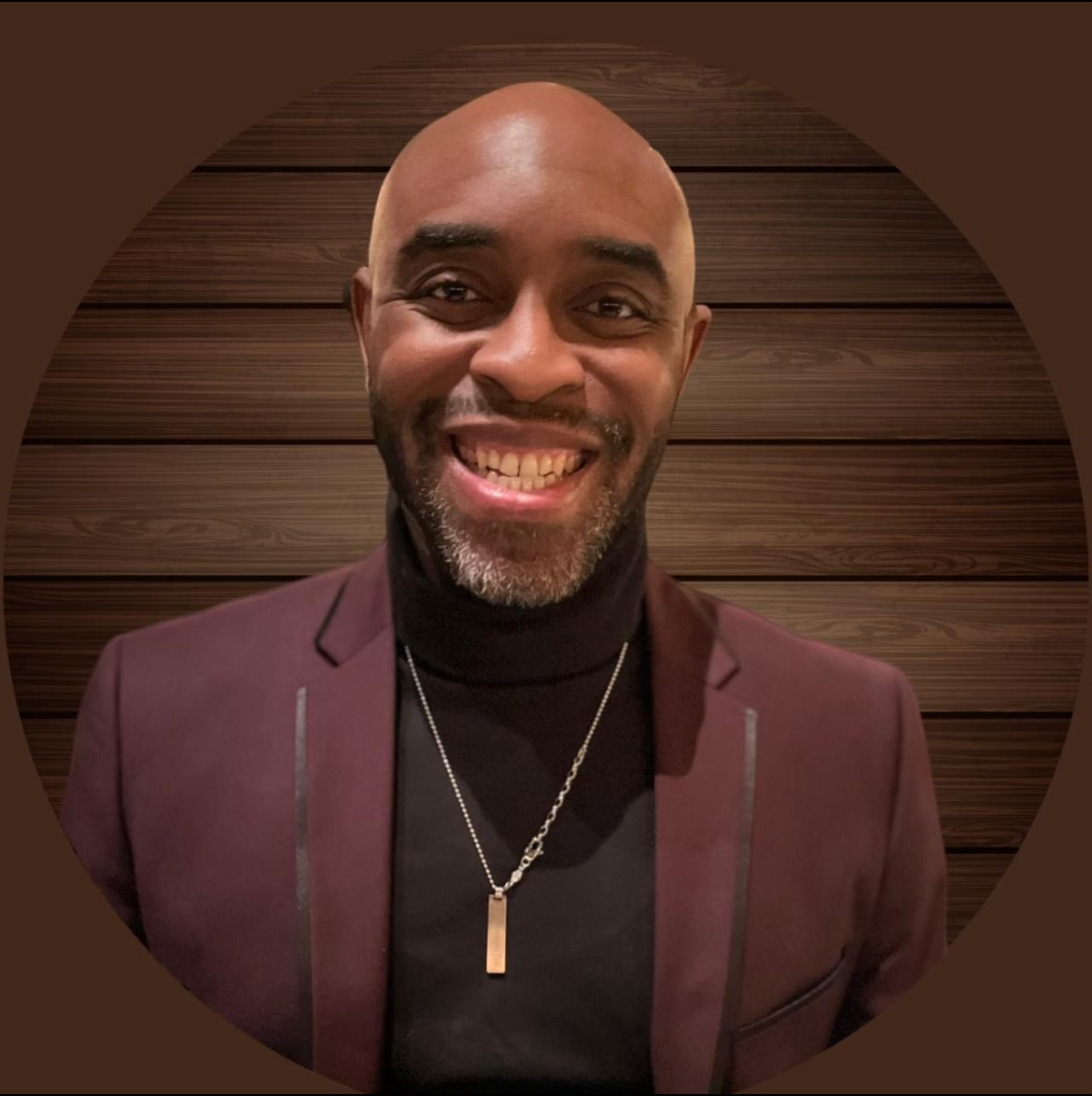 Smiling man in a maroon blazer and black turtleneck, wearing a silver necklace with a pendant, standing in front of a wooden background.