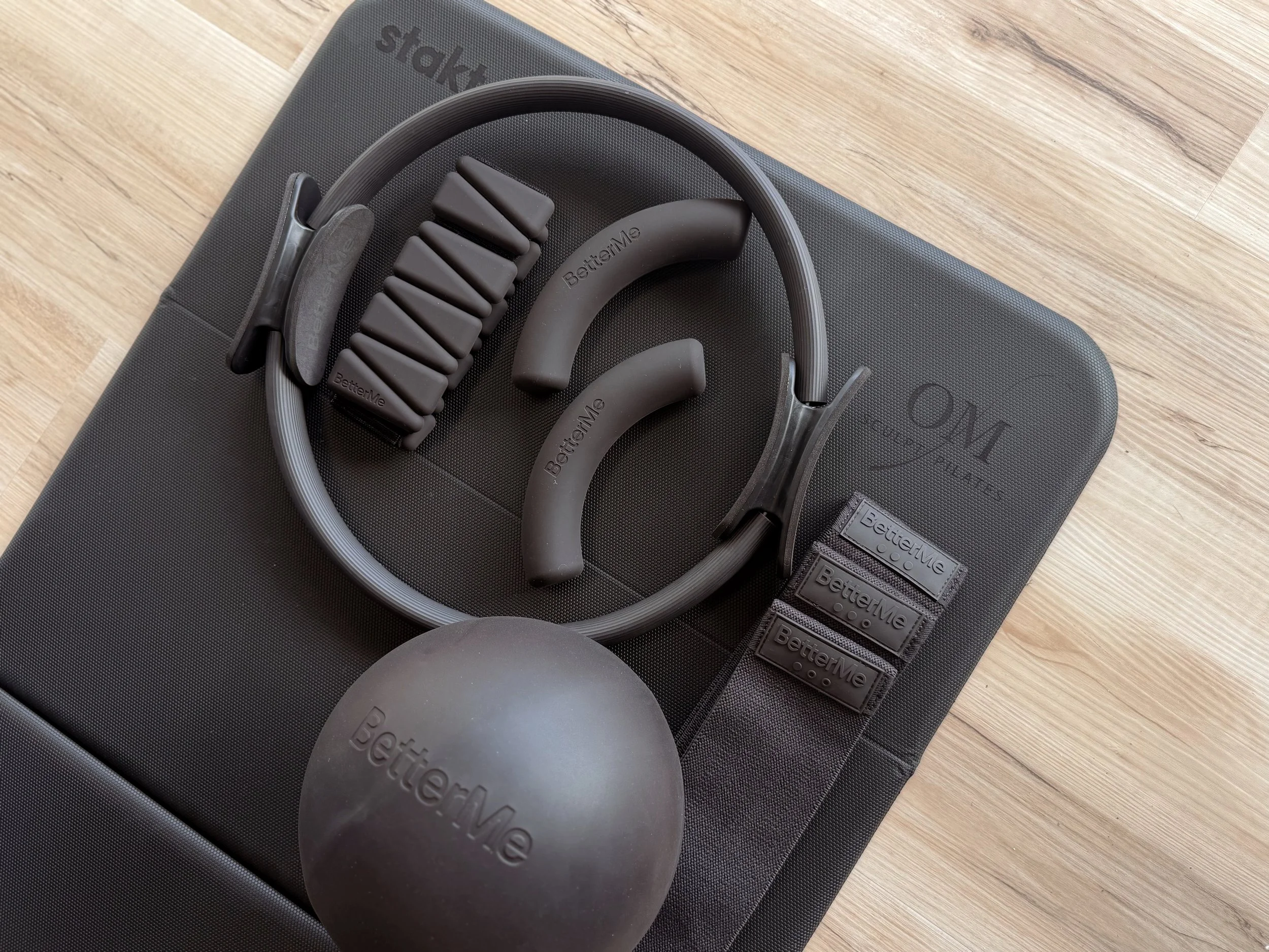 A set of black and gray fitness accessories including a weighted ball, resistance bands, and a head strap with padding, all arranged on a black fitness mat on a wooden floor.