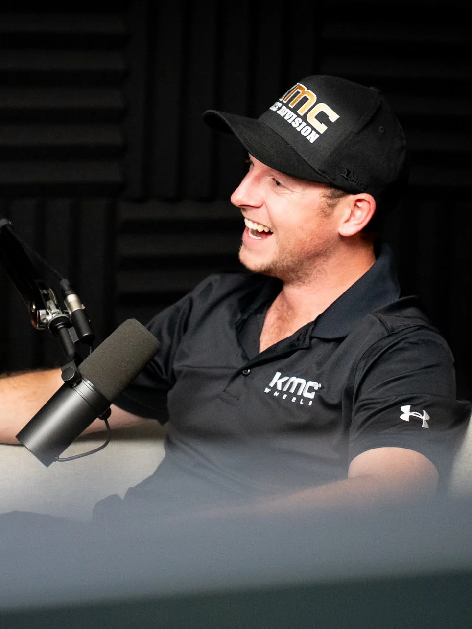 A man smiling and talking into a microphone in a recording studio, wearing a black cap and a black polo shirt with KMC logo.
