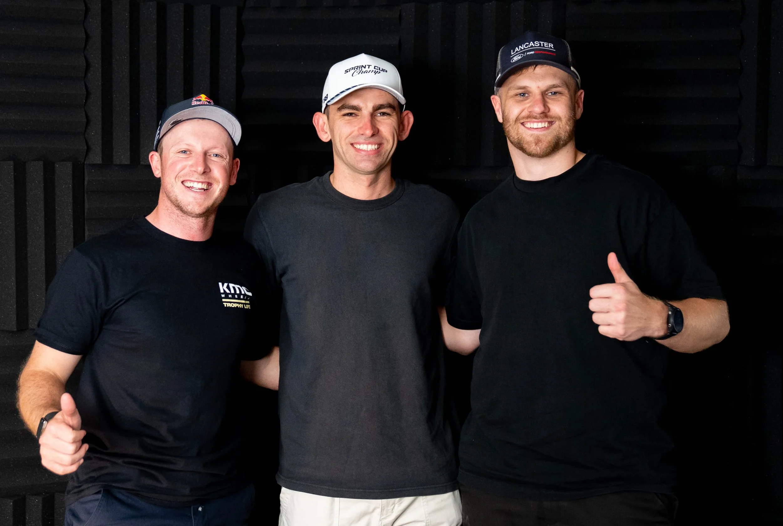 Three men standing close together in front of a black foam wall, smiling at the camera, wearing casual black t-shirts and hats.