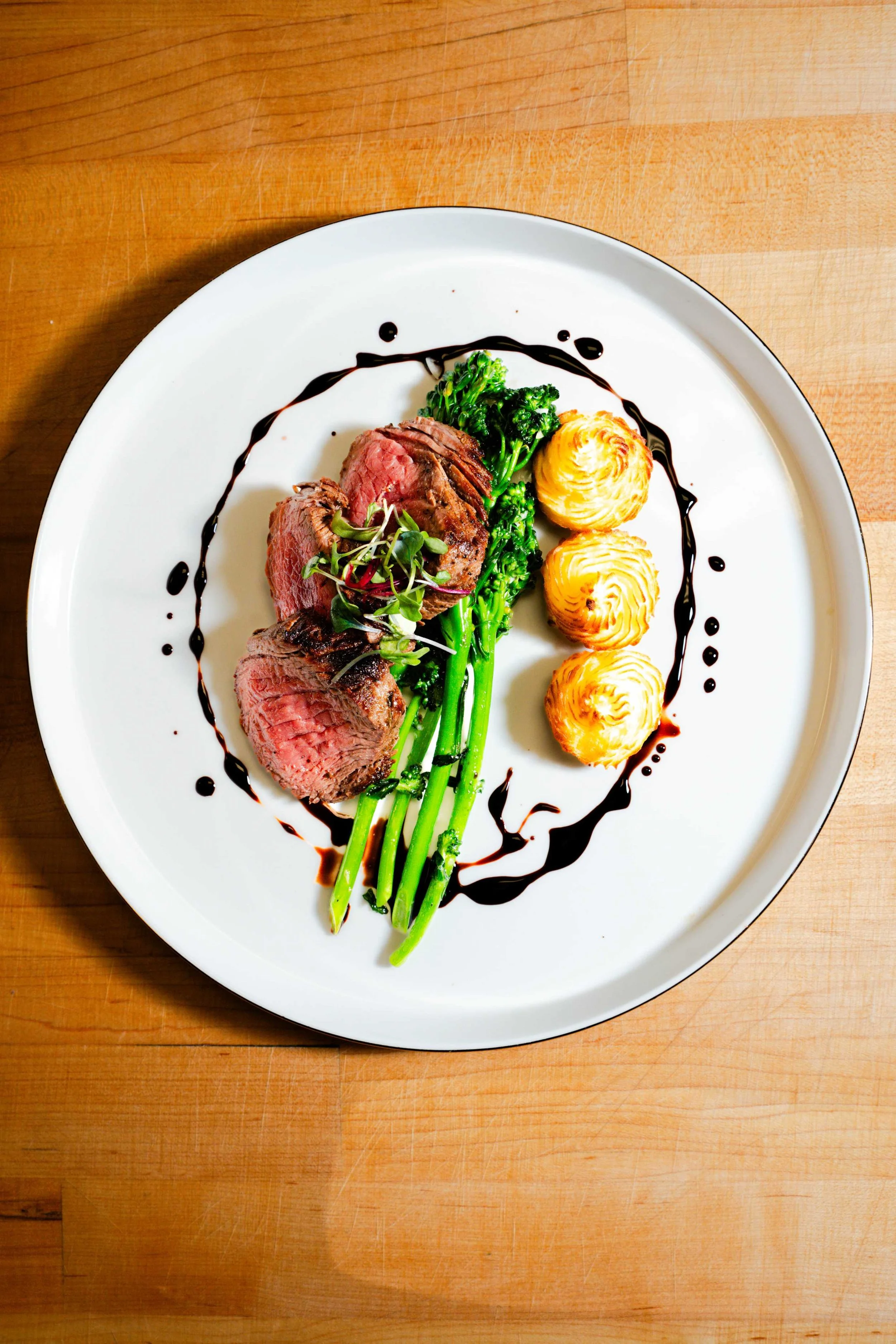 Plated steak medallions with broccolini, piped potatoes, microgreens, and balsamic reduction on a white plate.