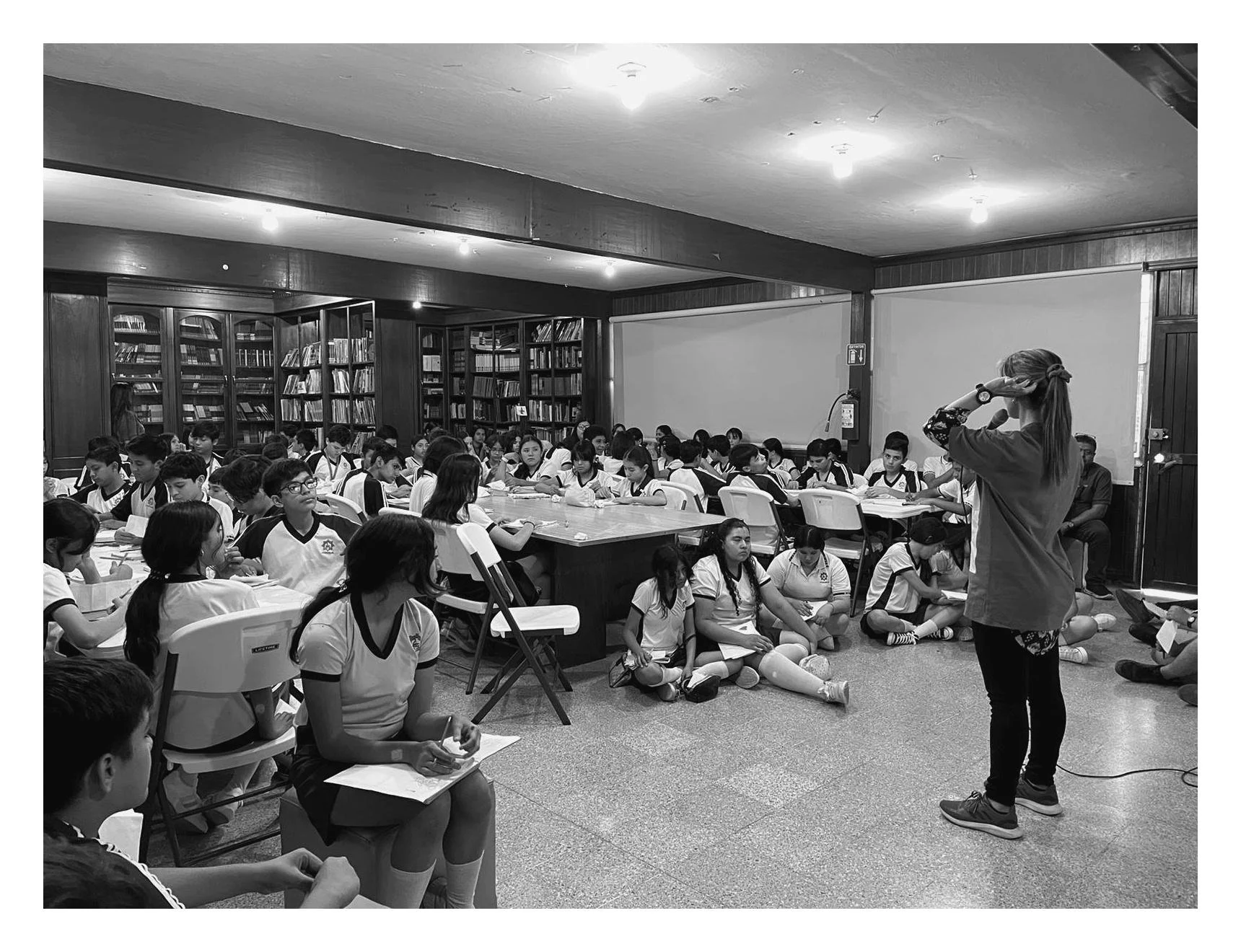 Clase o reunión en una biblioteca escolar con niños, algunos sentados en sillas y otros en el suelo, una mujer de pie da una charla, las estanterías llenas de libros y paredes de madera en el fondo.