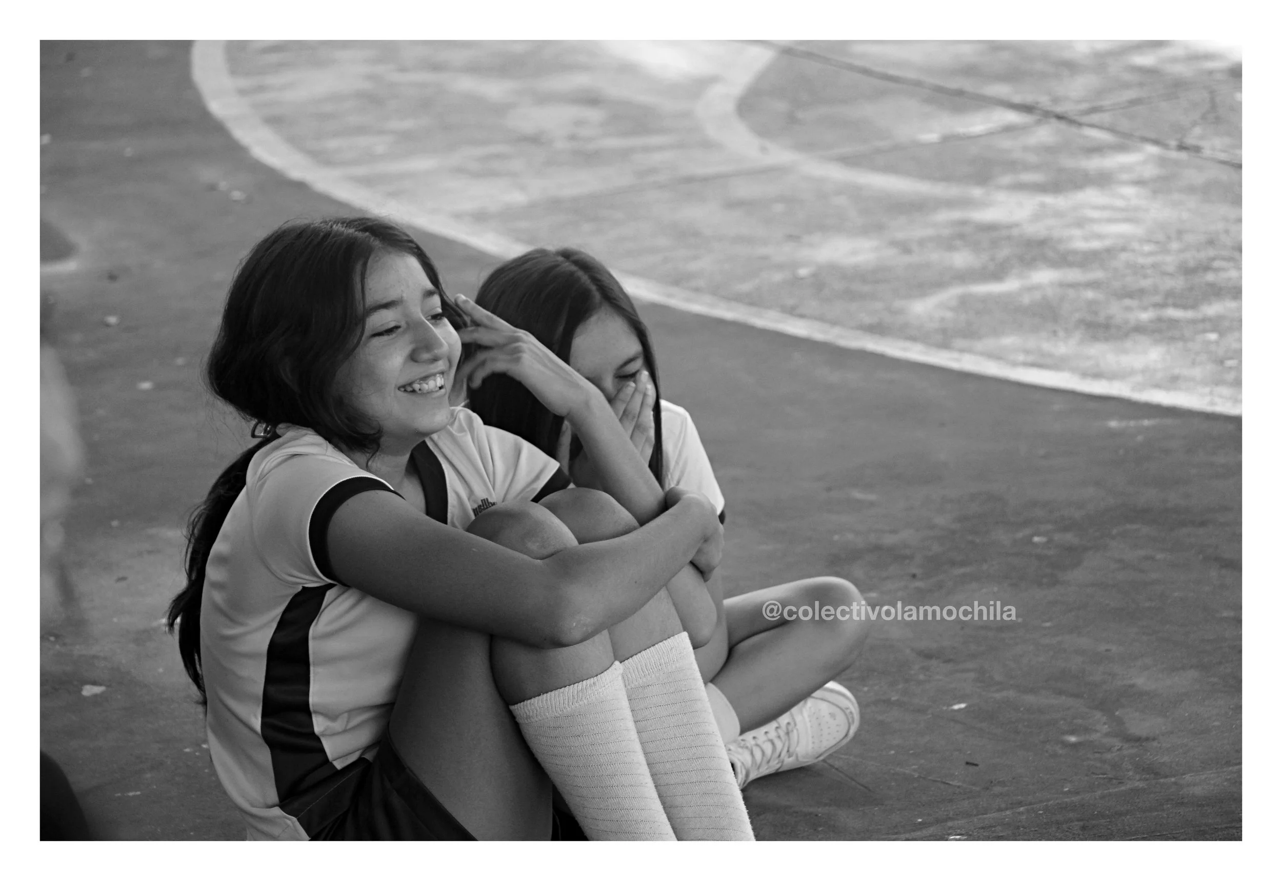 Dos niñas en uniforme de fútbol sentadas en el suelo, riendo y cubriéndose la cara, en una cancha de baloncesto o similar.