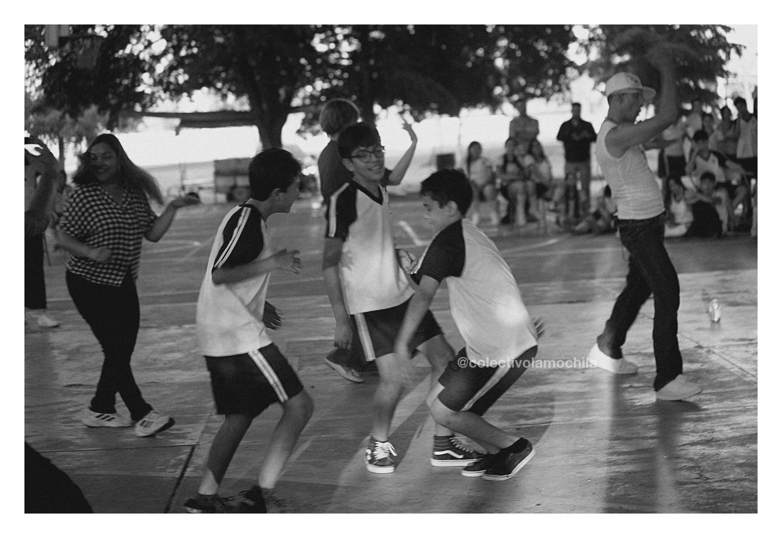 Niños jugando en una cancha cubierta, algunos bailando y otros saltando, con árboles y personas en el fondo