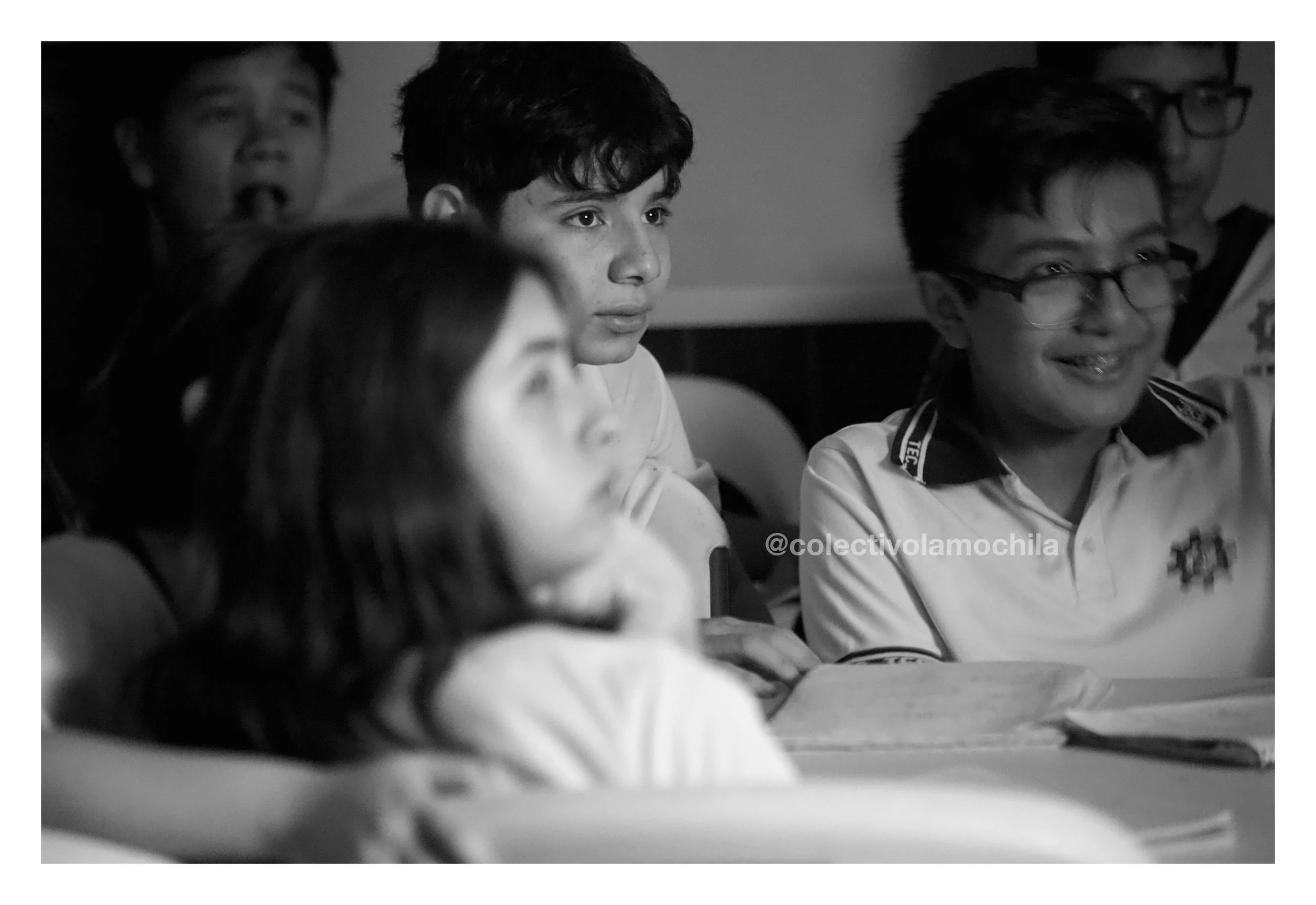 Grupo de niños en una clase, algunos observando y otros sonriendo. Uno lleva gafas y camiseta con logotipo, parece atento.