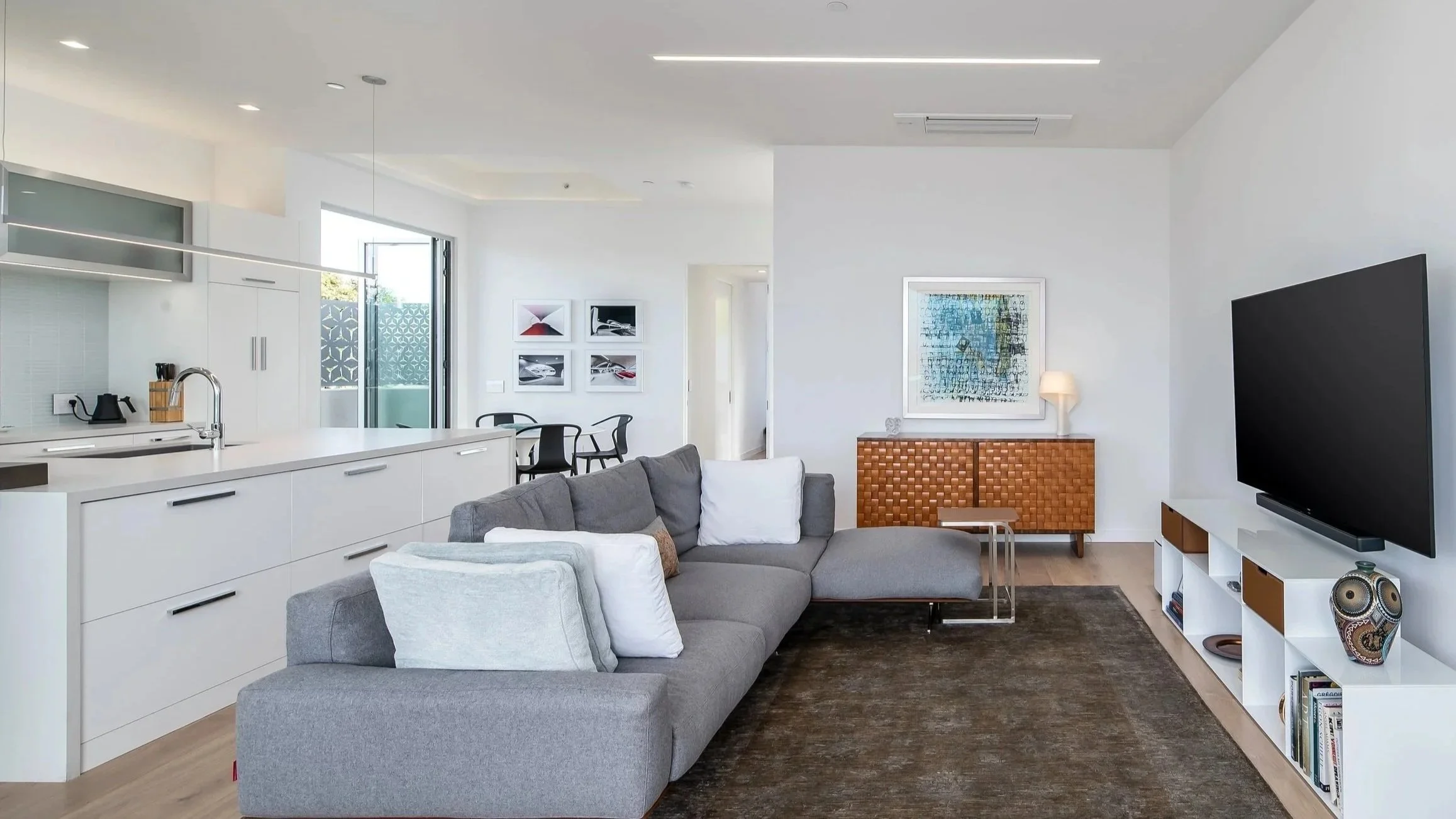 Modern living room with white walls, a gray sectional sofa, a flat-screen TV on a white stand, framed artwork on the walls, a wooden sideboard, and a dark rug on light wooden flooring.