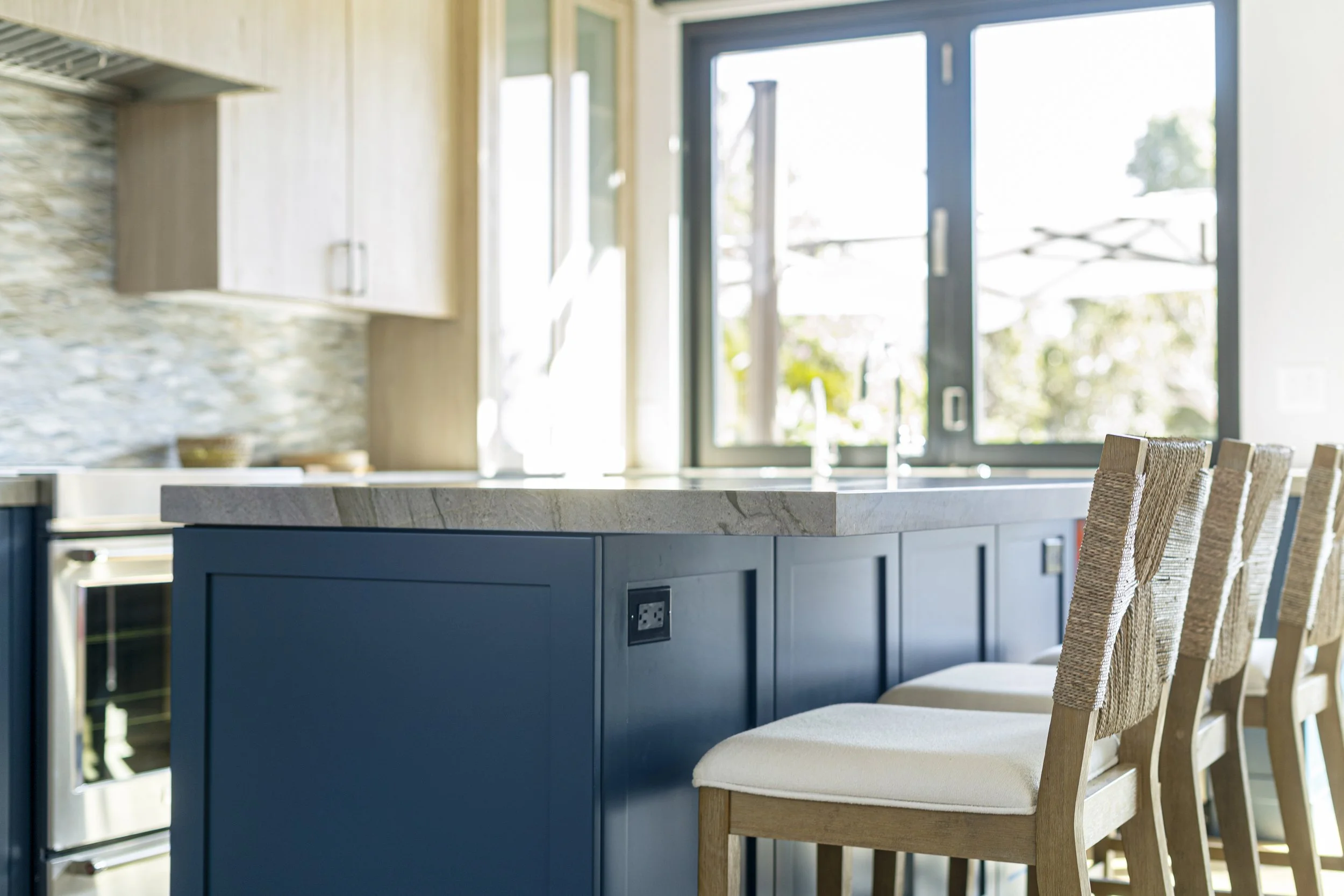 Modern kitchen with a blue island, beige chairs with woven backs, through large sliding glass doors with an outdoor view.