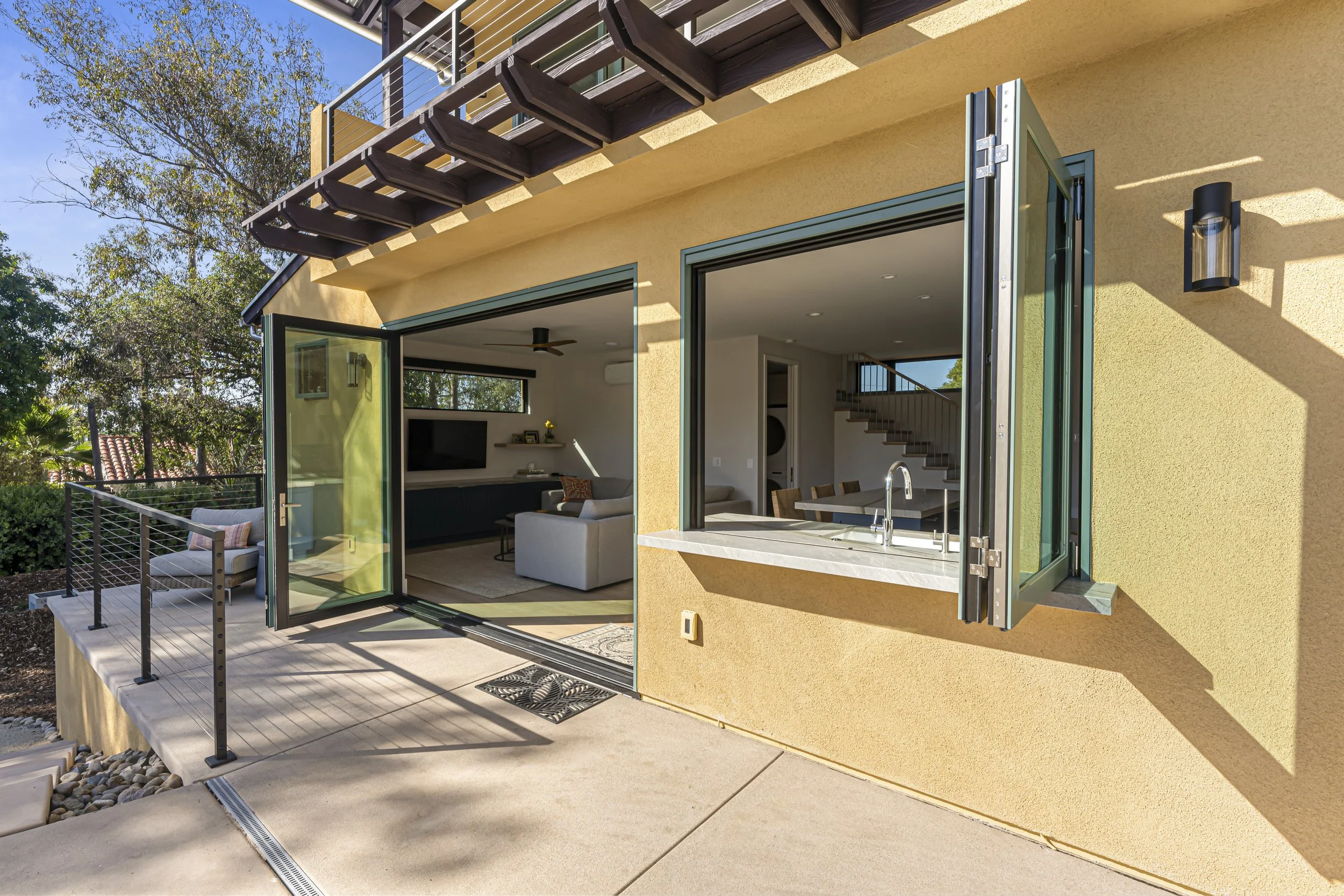 View of a modern house patio with open glass doors leading inside, outdoor seating, and a balcony above in a sunny, suburban setting with trees.