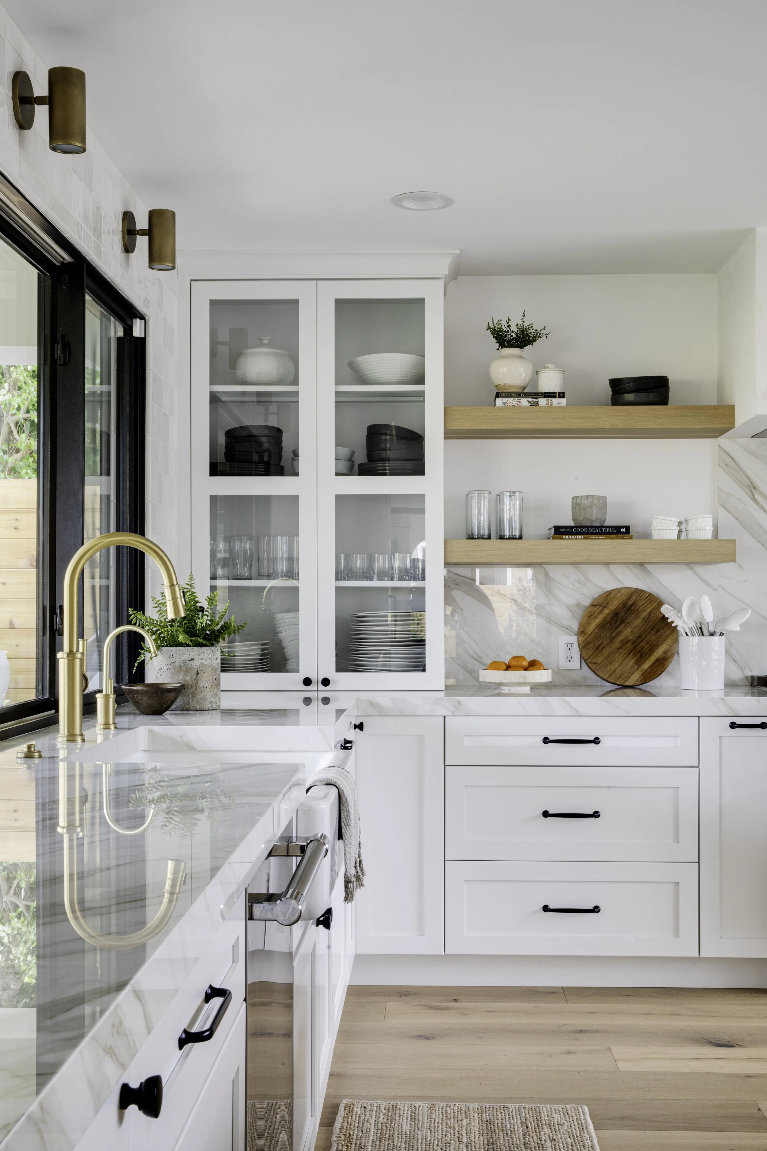 Modern kitchen with white cabinets, marble countertops, gold faucet, glass-front cabinet filled with dishes, open wooden shelves with decorative items, and a window with natural light.