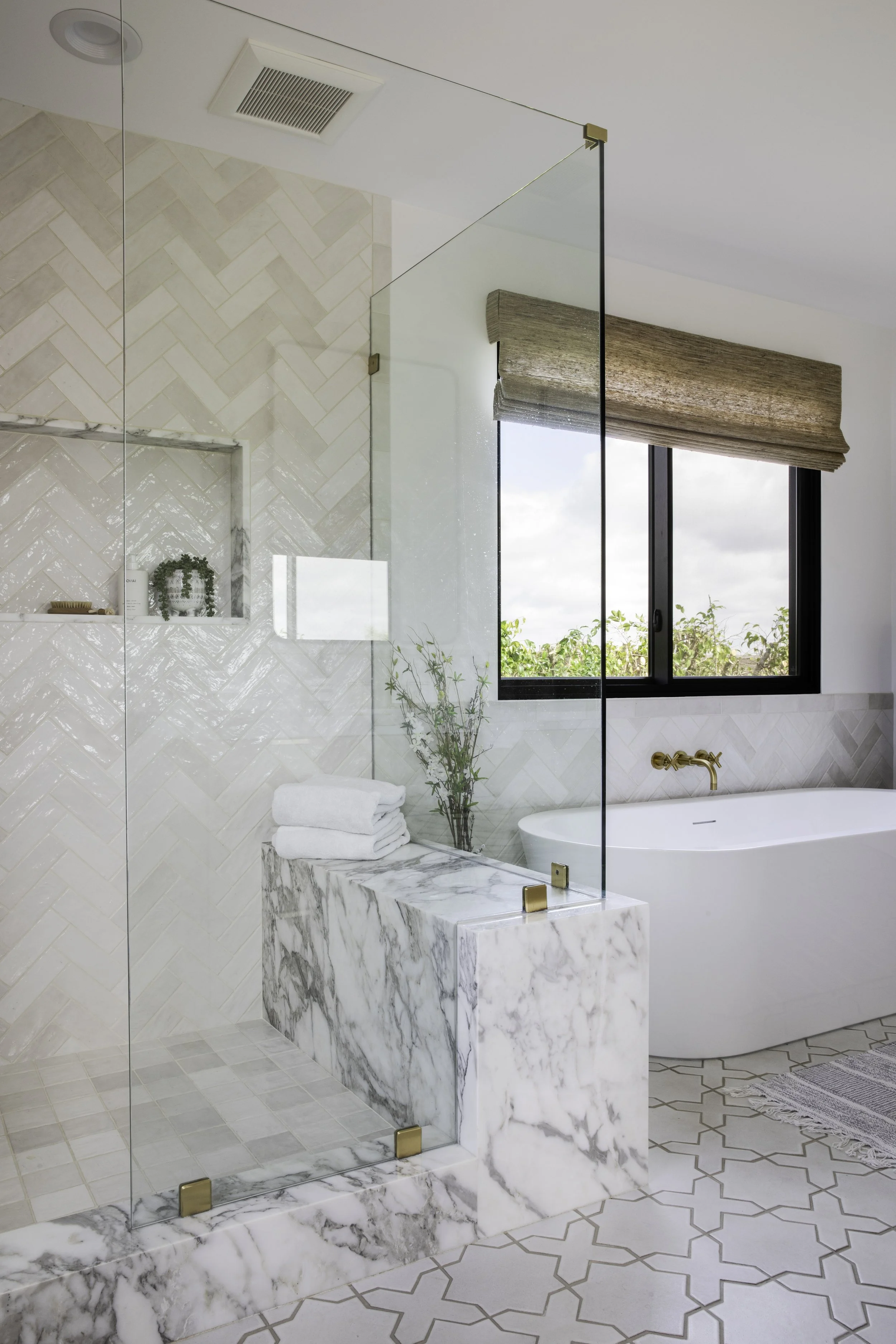 Modern bathroom with a glass shower door, a marble countertop, a large window with a beige Roman shade, and a rolled towel on the countertop.
