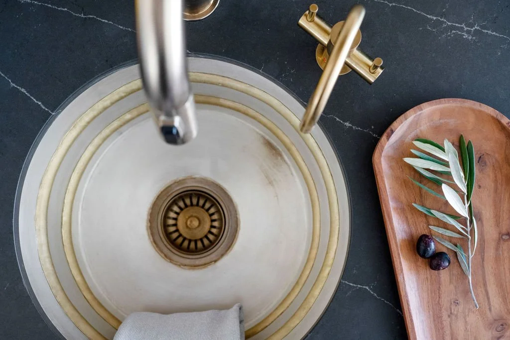 Top-down view of a kitchen sink with a stainless steel faucet, a brass faucet, and a wooden tray with green leaves and two purple figs on a dark countertop.
