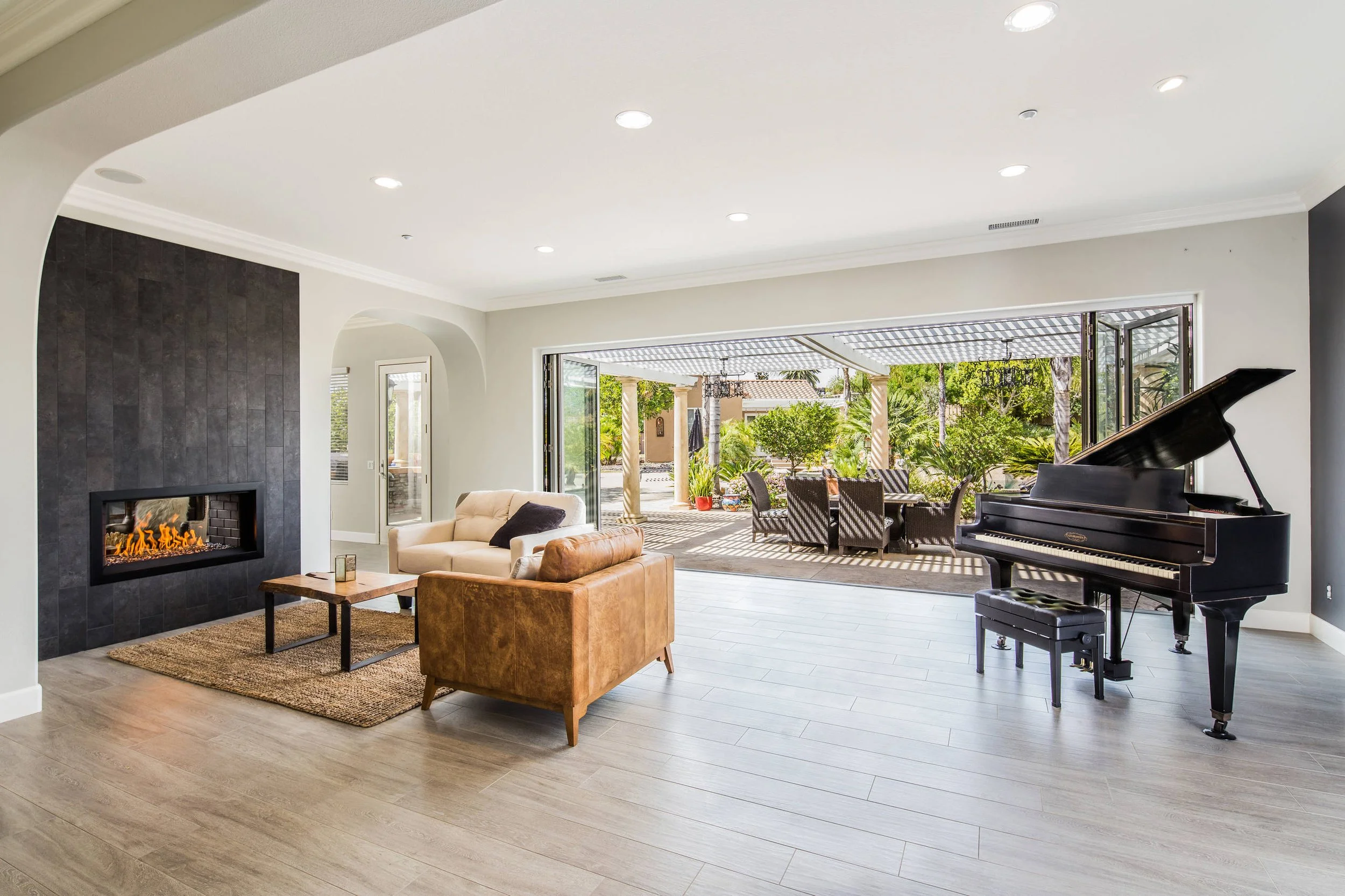 A spacious living room with a black grand piano, a beige sofa, a brown leather armchair, a rectangular coffee table, a fireplace with a dark gray tiled wall, and large open sliding glass doors leading to an outdoor patio with trees and outdoor furnit