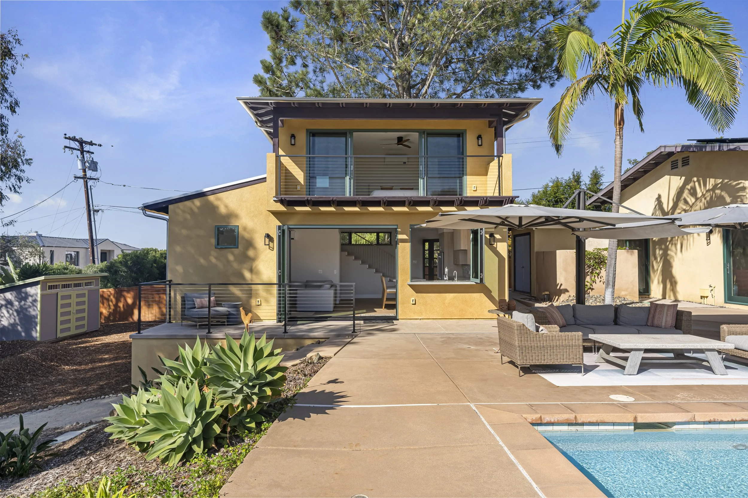 Front view of modern two-story yellow house with outdoor living space, patio, and pool. There are patio furniture, parasol, lush greenery, and trees, with a blue sky overhead.
