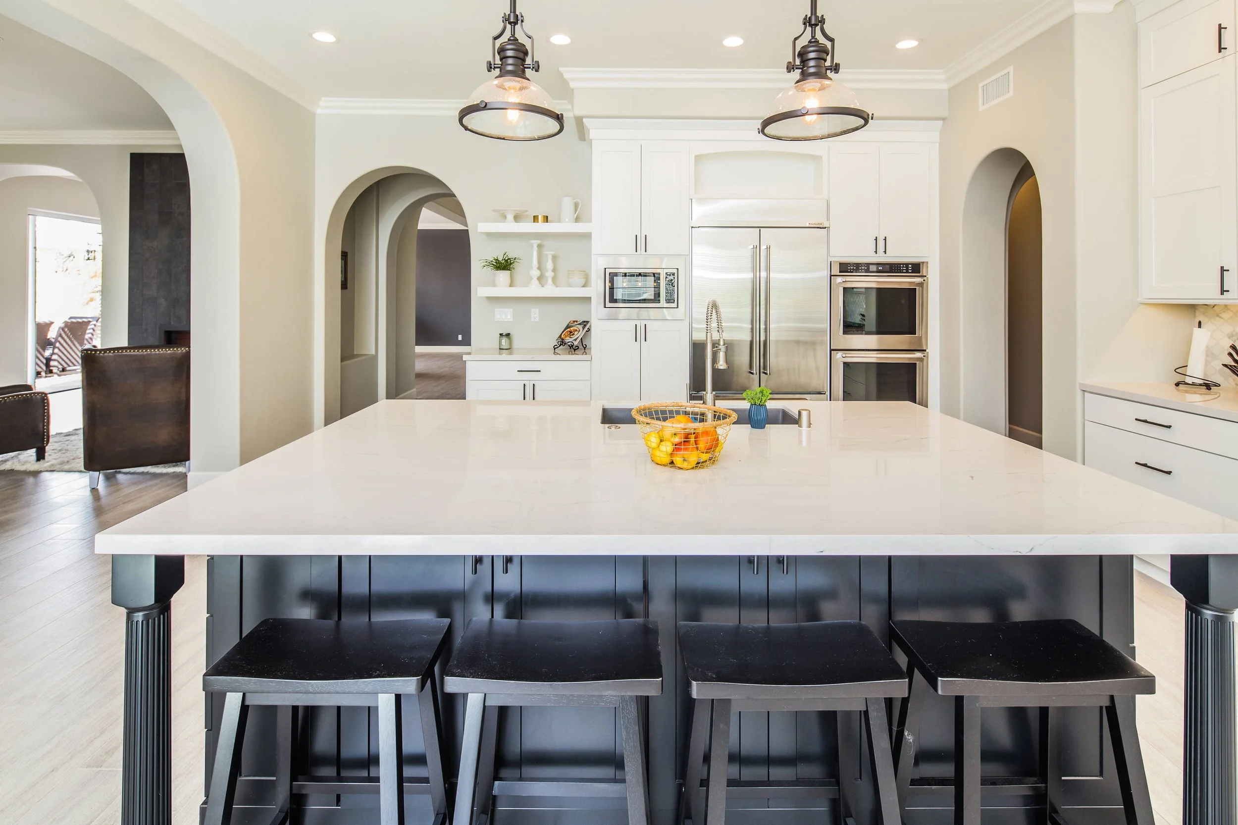 Modern kitchen with a large central island, black chairs, white cabinets, stainless steel appliances, and hanging light fixtures.