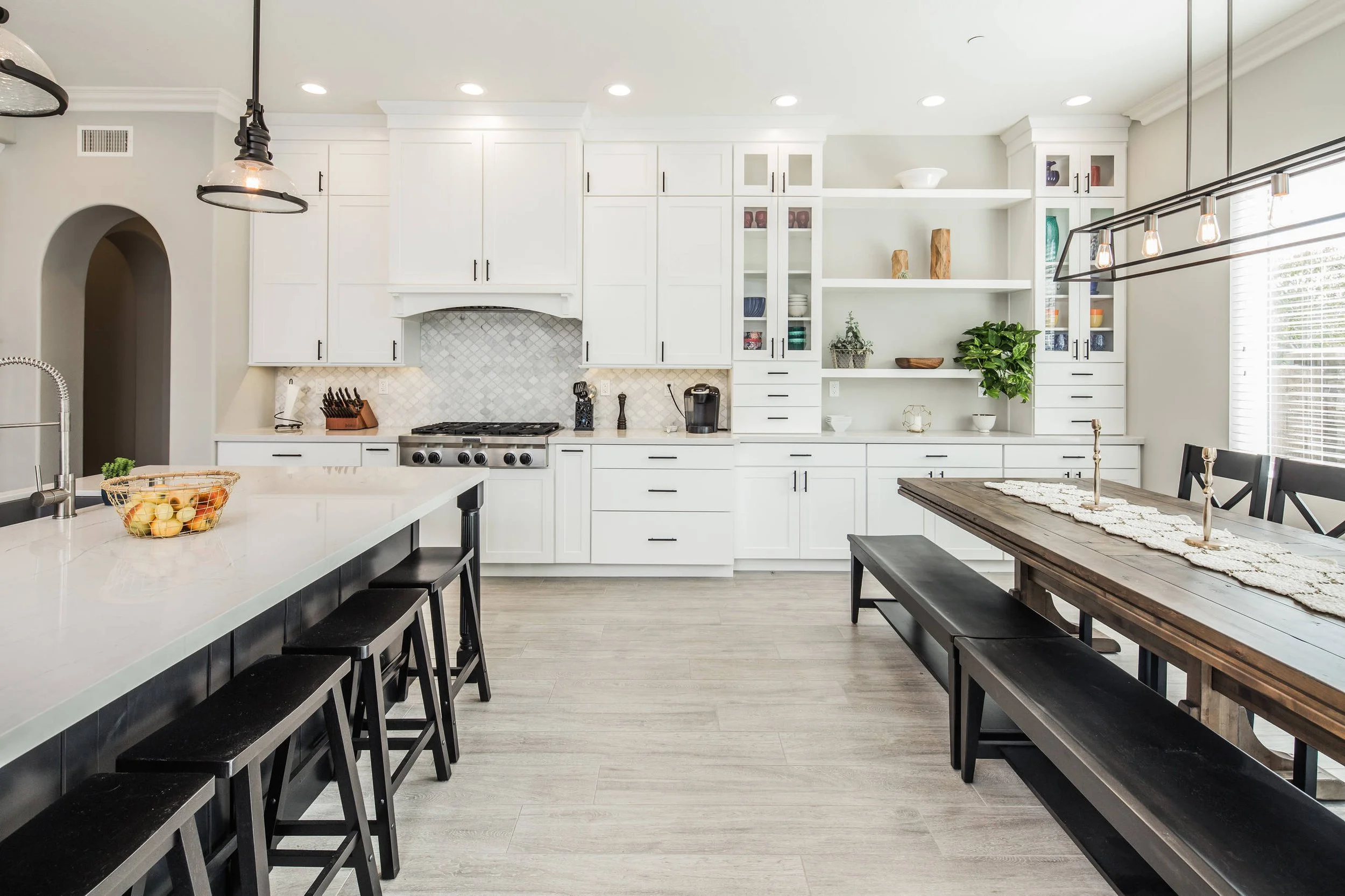 Modern kitchen with white cabinets, a large island with black stools, open shelves with decorative items, a dining table with a runner and candelabras, pendant lighting, and a window with blinds.