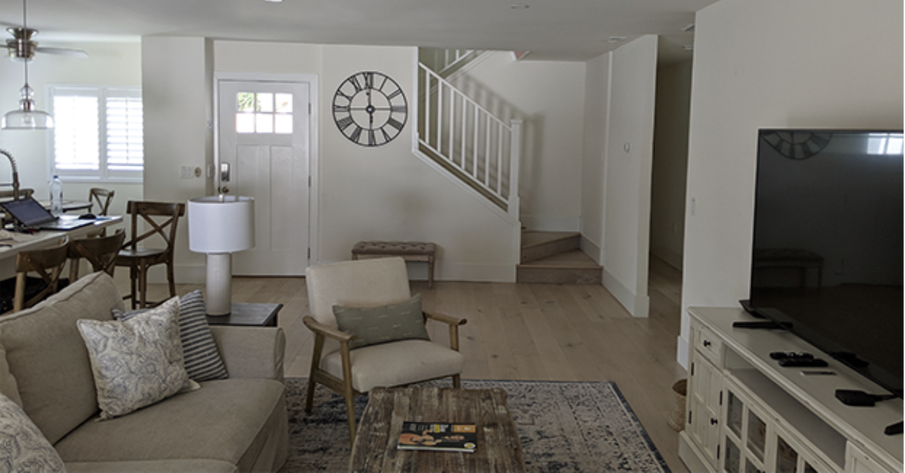 Living room with beige sofa, armchair, and wooden coffee table, TV on white stand, and dining area with wooden chairs and table in background. Wall clock and staircase are visible.