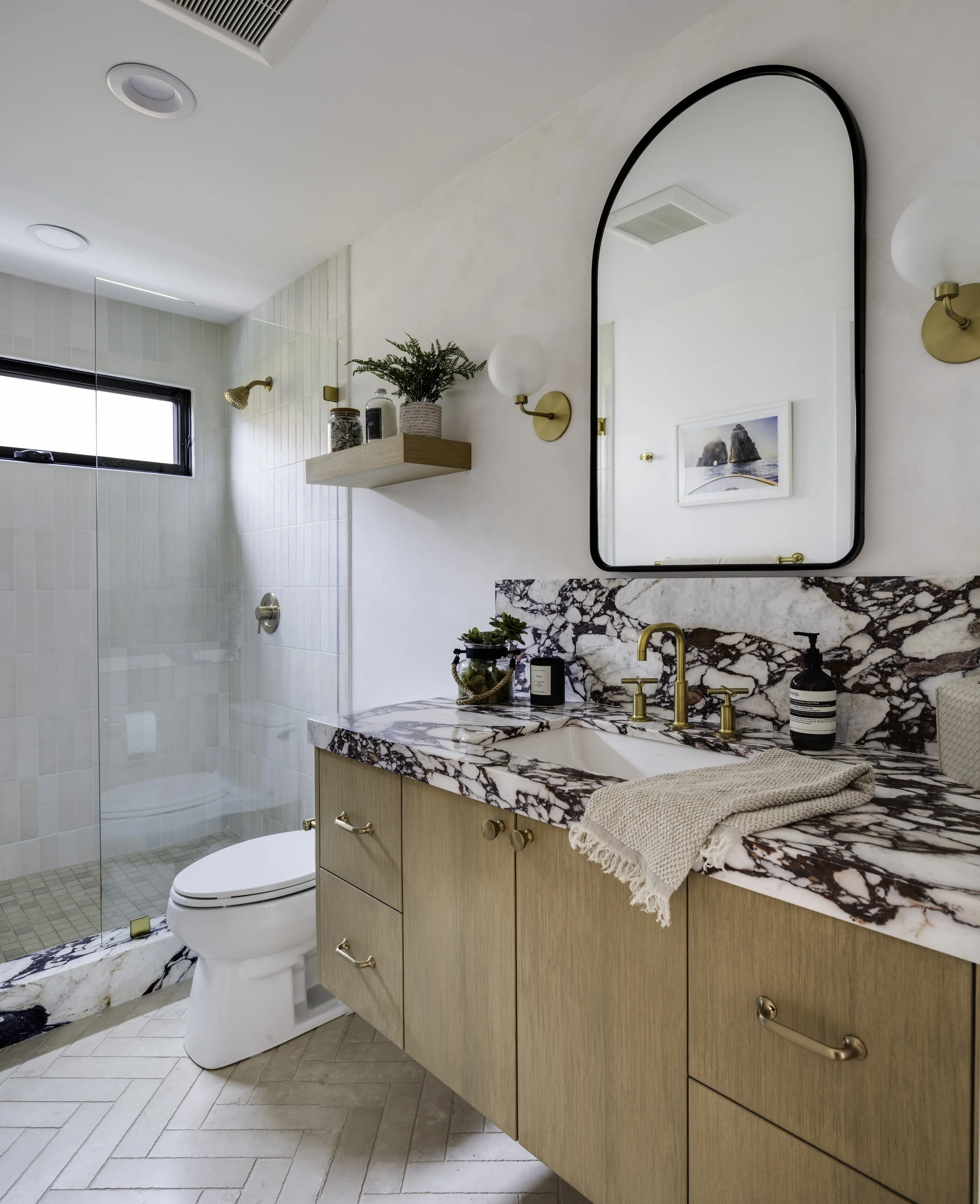 Modern bathroom with a marble countertop vanity, brass fixtures, a large wall mirror, a potted plant on a floating shelf, a glass-enclosed shower with white tiled walls, a window, a towel, and decorative elements.