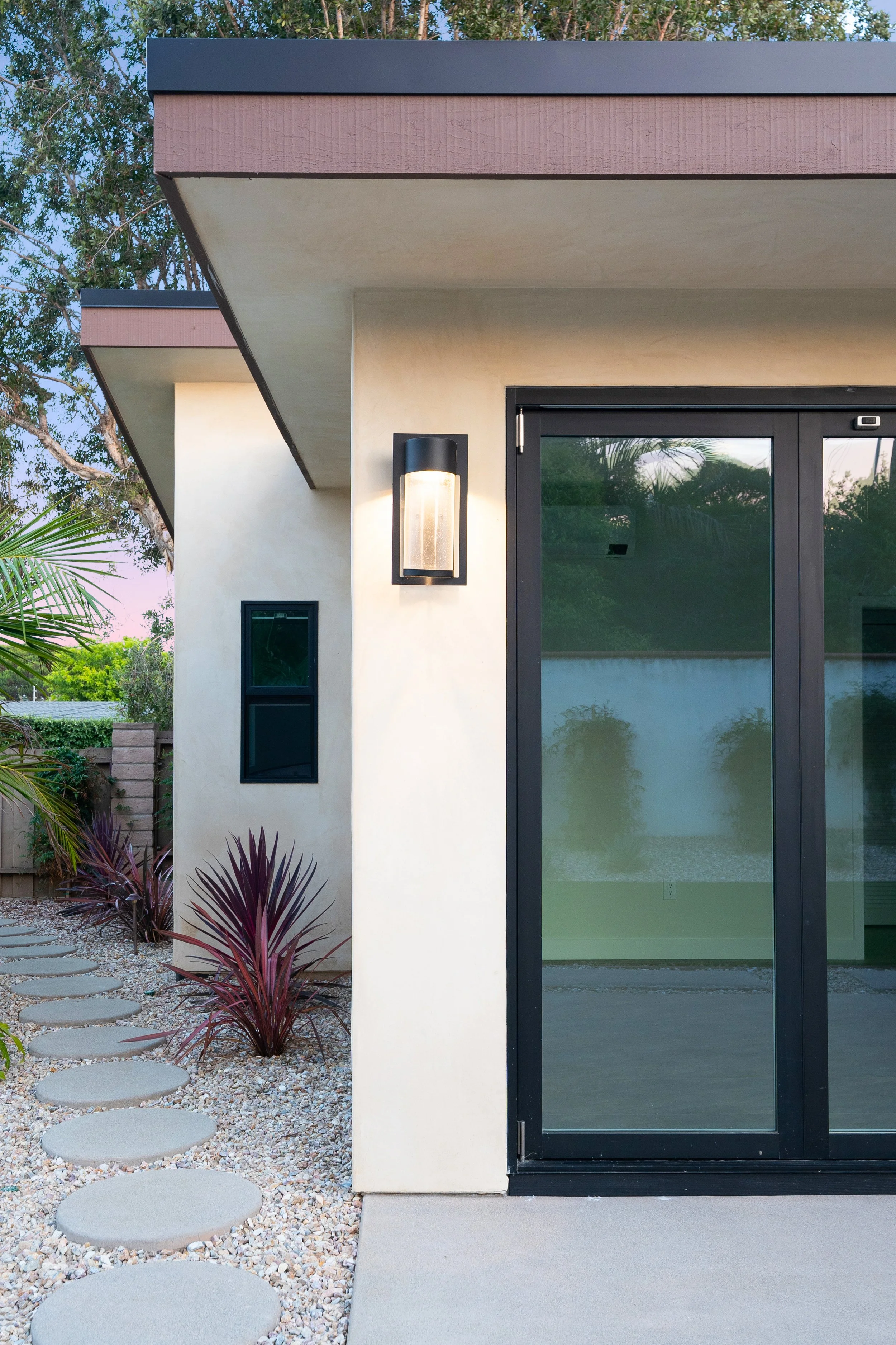 Modern house exterior with beige walls, black-framed glass sliding door, exterior wall sconce, and landscaped yard with stepping stones and purple plants.