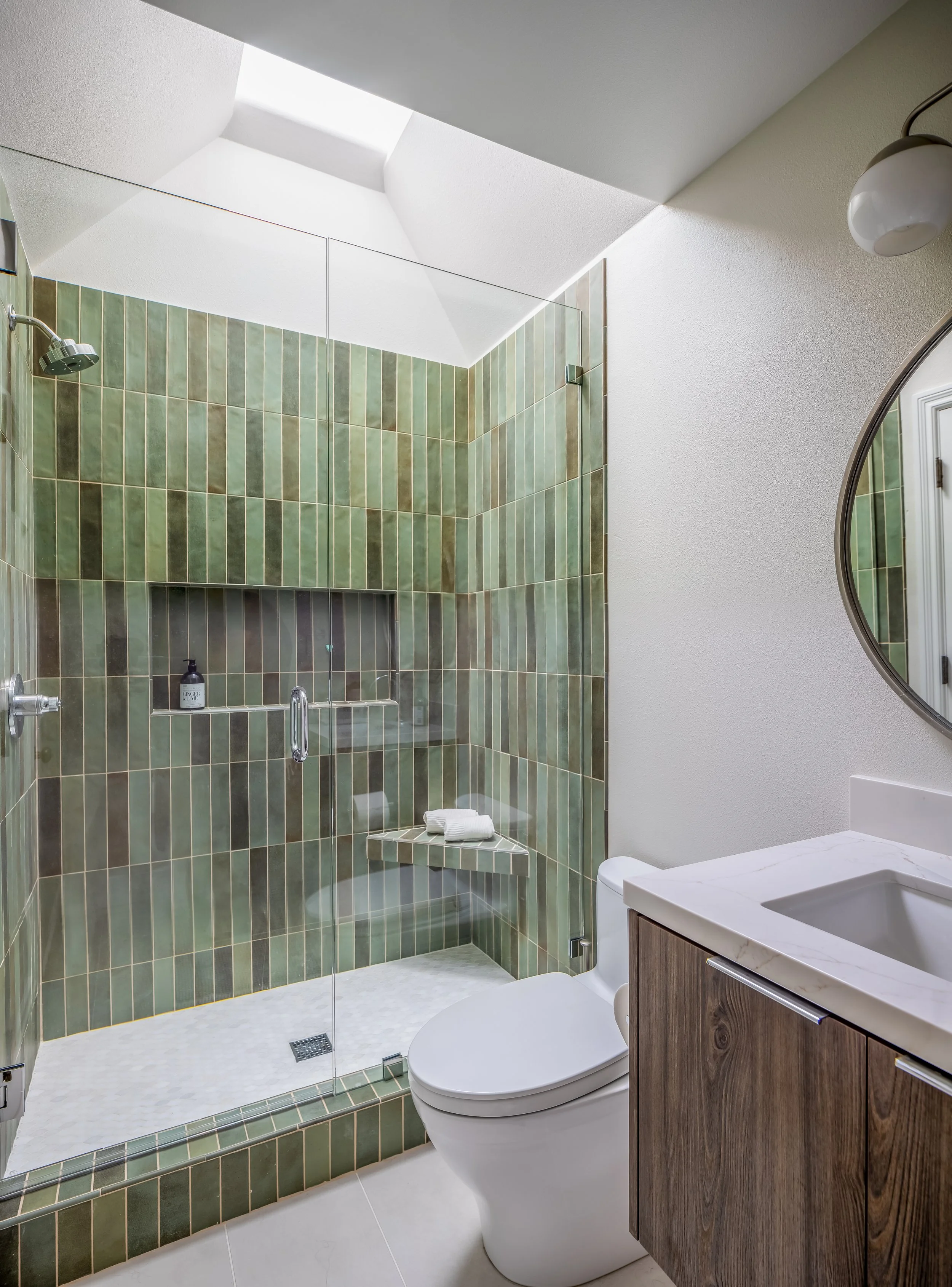 Modern bathroom with walk-in glass shower, green tiled walls, white ceiling with skylight, wooden vanity with sink, toilet, and round mirror.