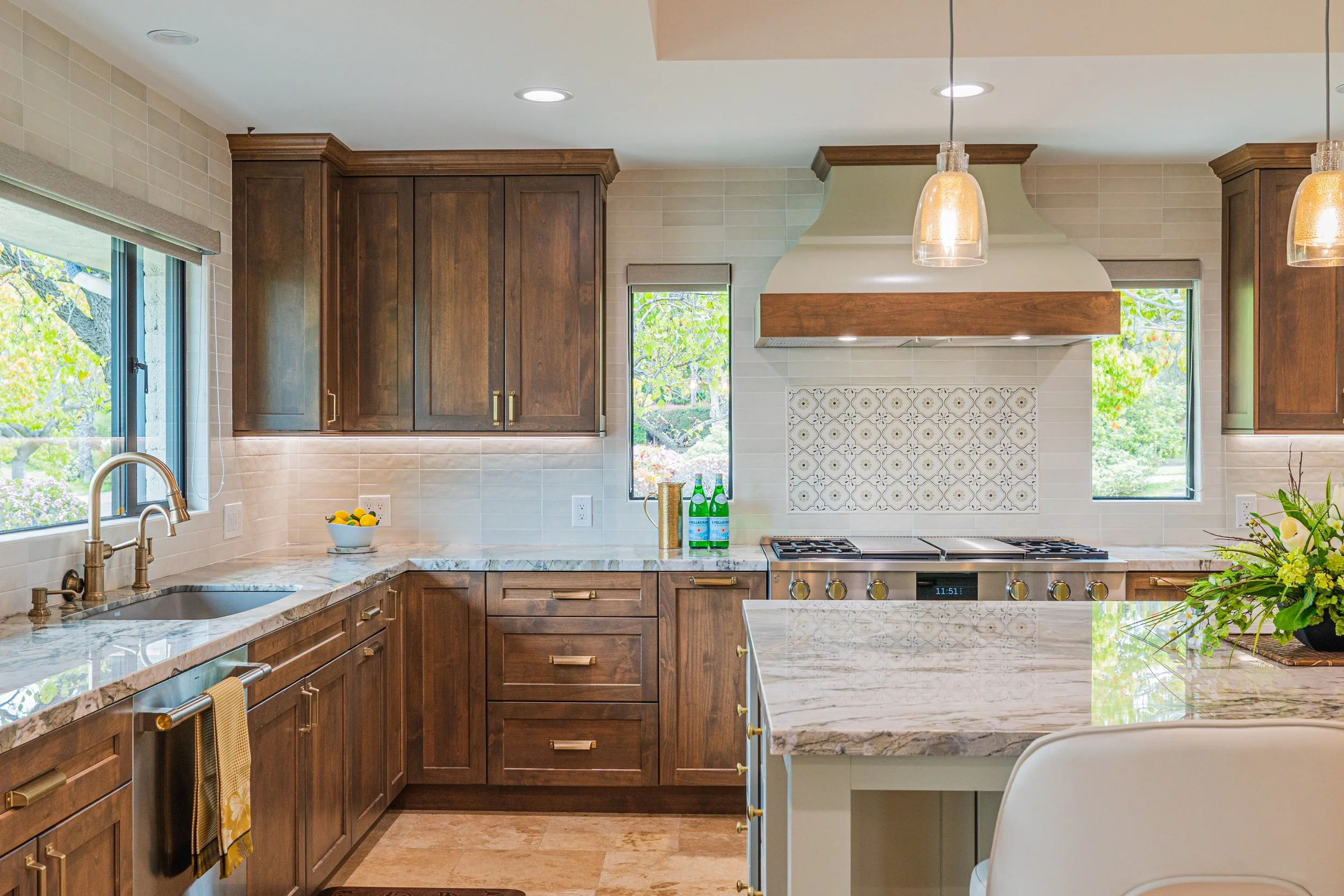 A modern kitchen with wooden cabinets, marble countertops, a stainless steel stove, large windows showing greenery outside, pendant lights hanging from the ceiling, and a bowl of lemons on the counter.