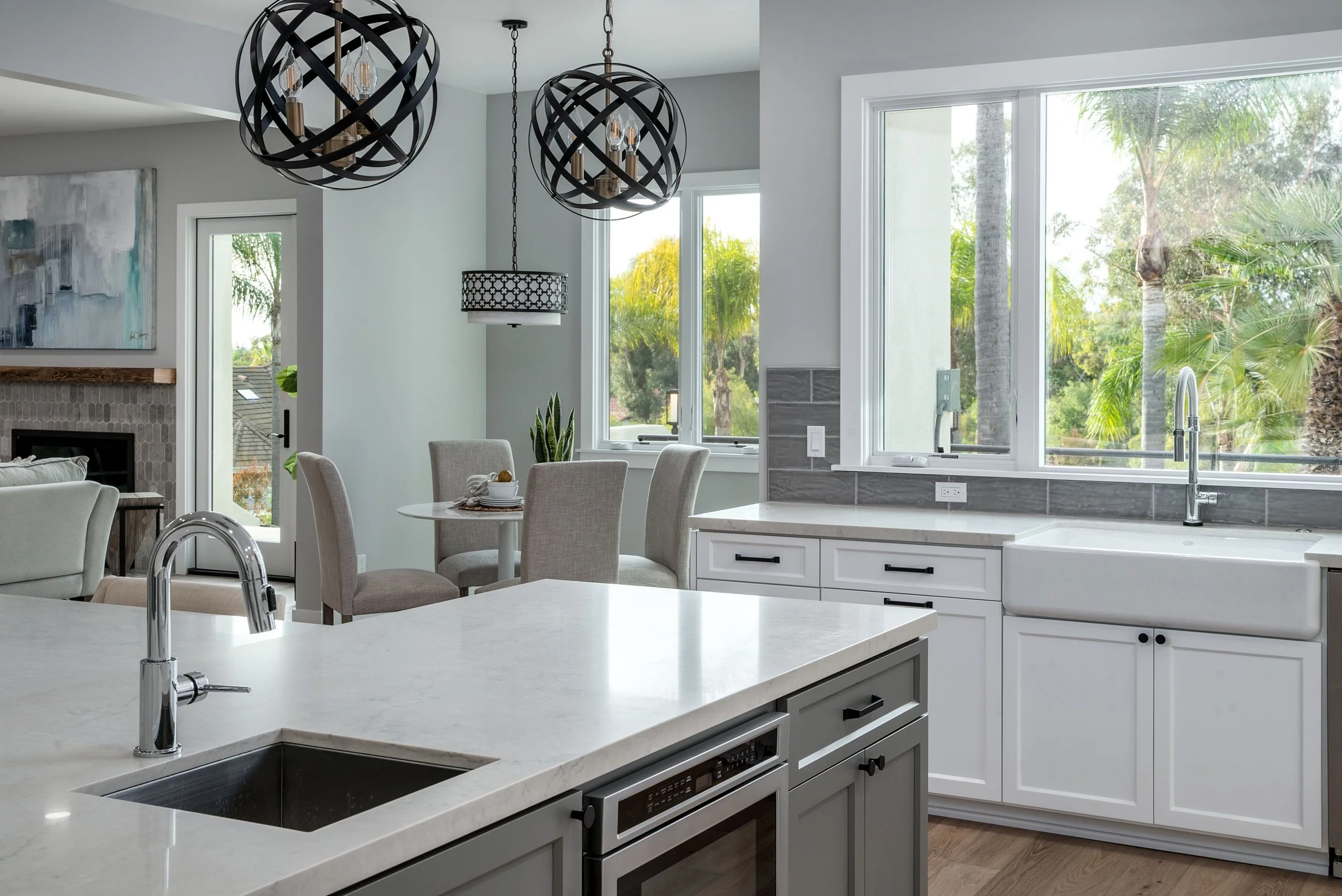 Modern kitchen with white cabinets, marble countertops, large windows showing greenery outside, black and gold chandeliers, and a dining area with beige chairs.