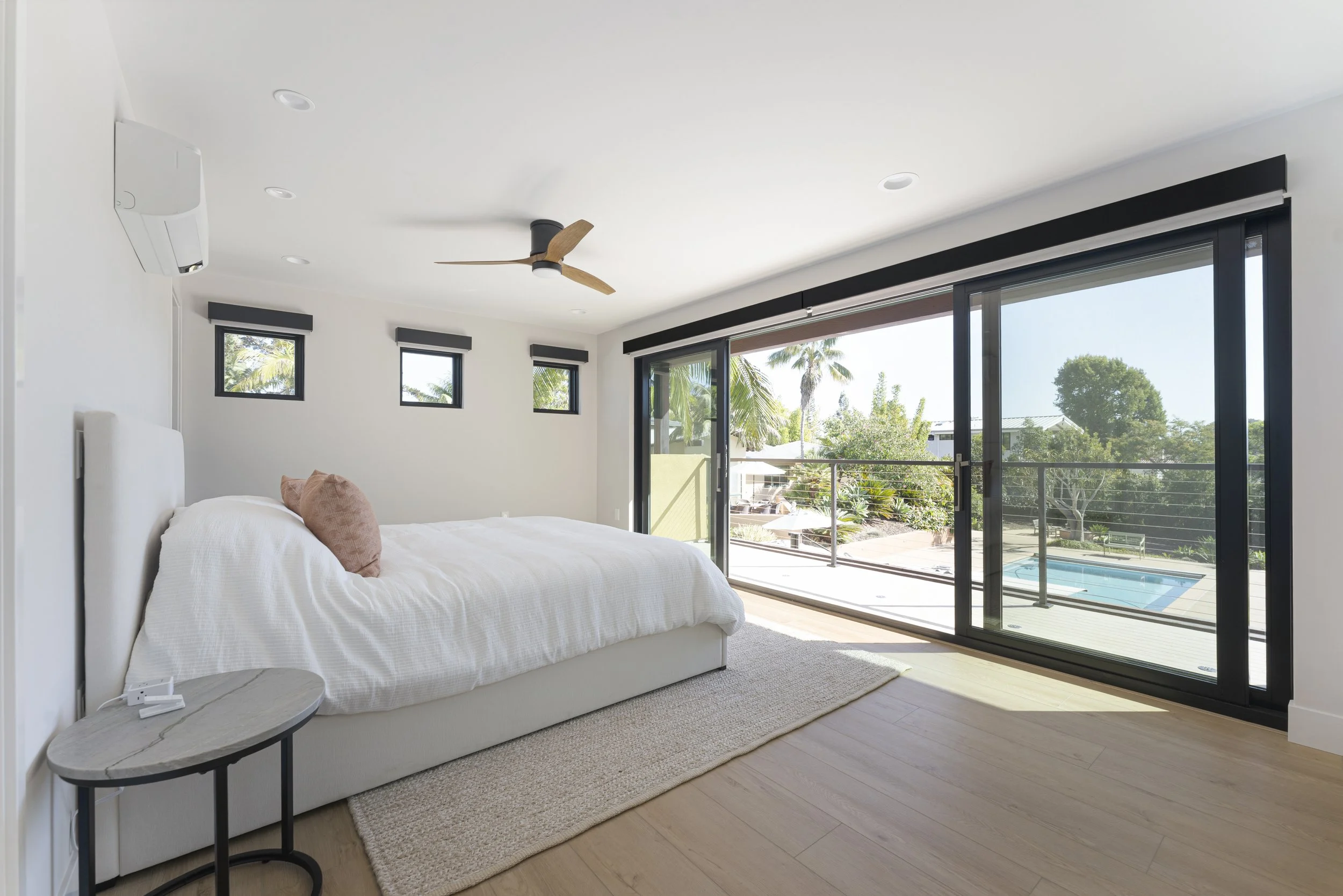 Modern bedroom with large sliding glass doors opening to a balcony view of trees, a pool, and a sunny sky. The room features white walls, a white bed with a pink pillow, small black window valances, a ceiling fan with wooden blades, and a small round