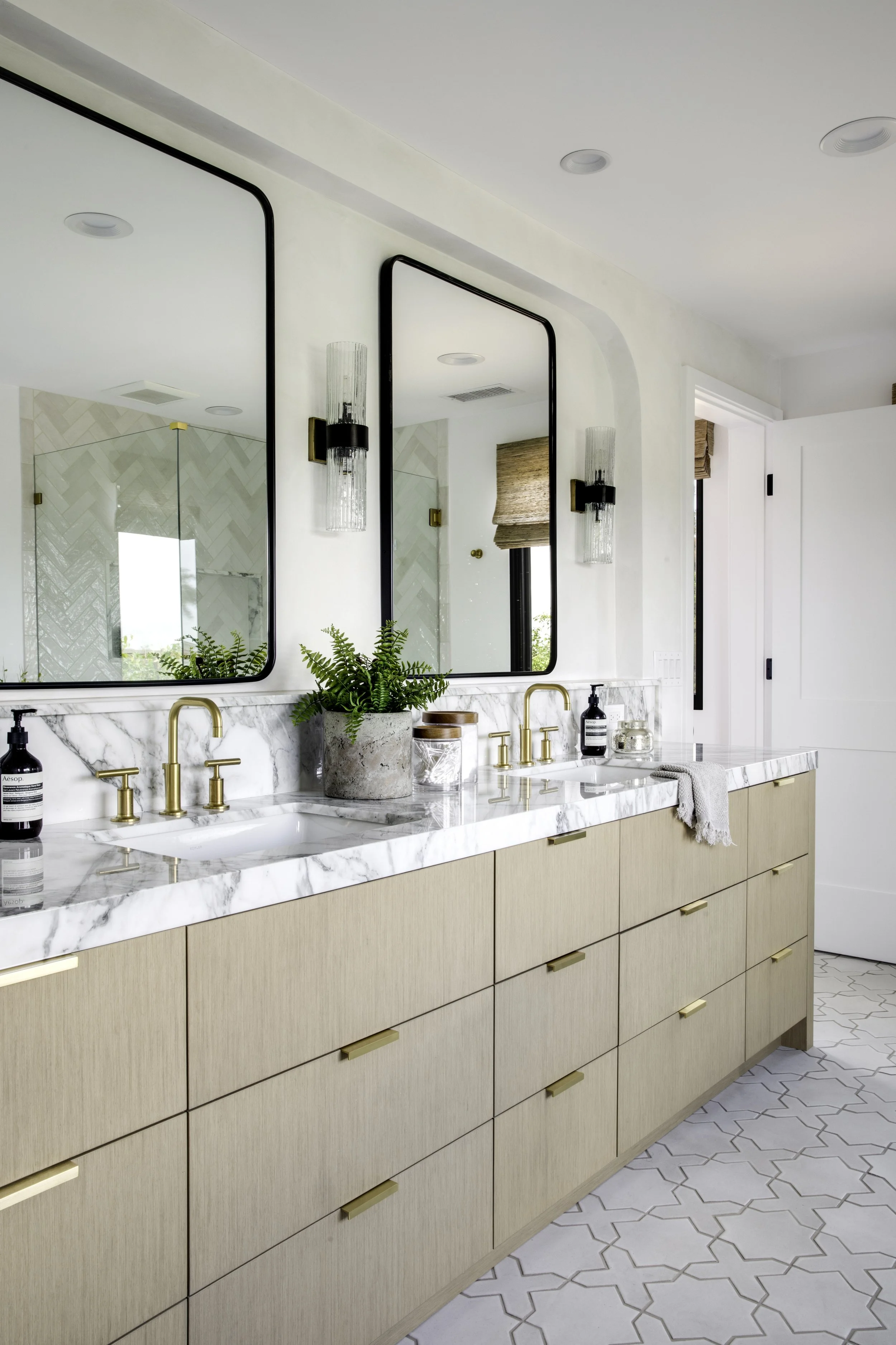 Modern bathroom with beige cabinets, marble countertop, gold fixtures, large mirrors, wall sconces, and patterned tile flooring.