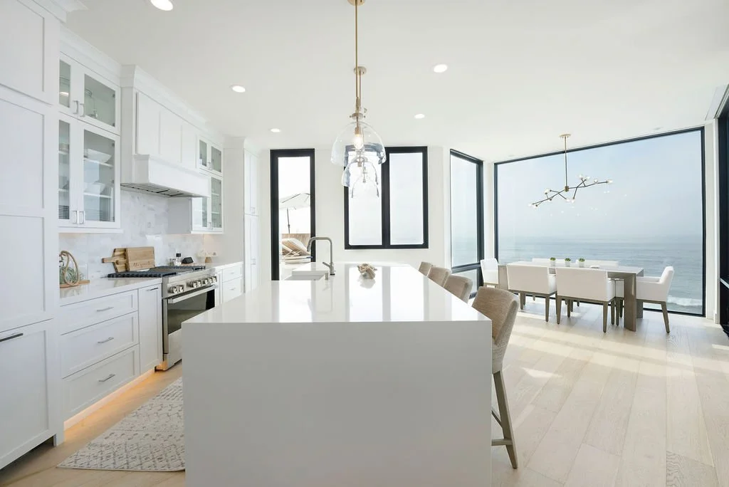 Modern kitchen and dining area with large floor-to-ceiling windows overlooking the ocean, white cabinets, island counter, and dining table with chairs.