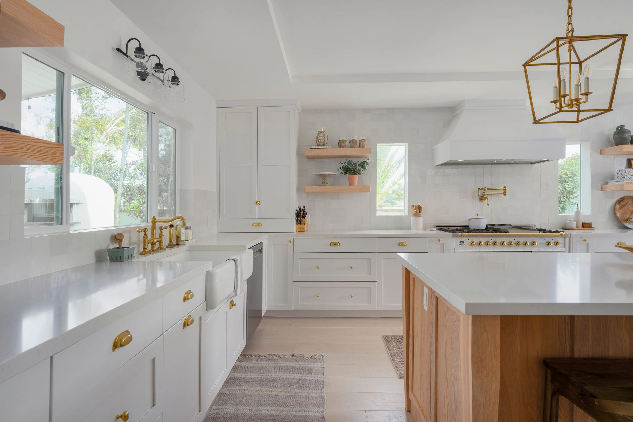 Bright kitchen with white cabinets, gold hardware, a white countertop, and wooden accents. Large windows with greenery outside, gold chandelier, and open shelving with decorative items.