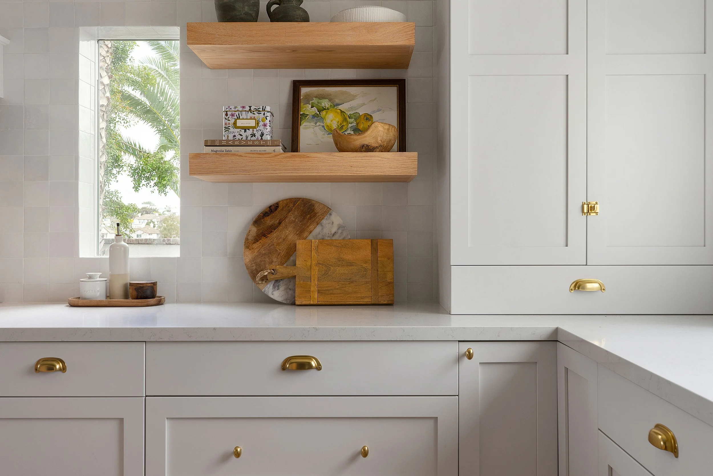 A kitchen countertop with a white finish, decorated with a tray holding a white bottle, a small container, and a wooden bowl, beneath wooden floating shelves holding books, artwork, a wooden bowl with fruit, and decorative jars.