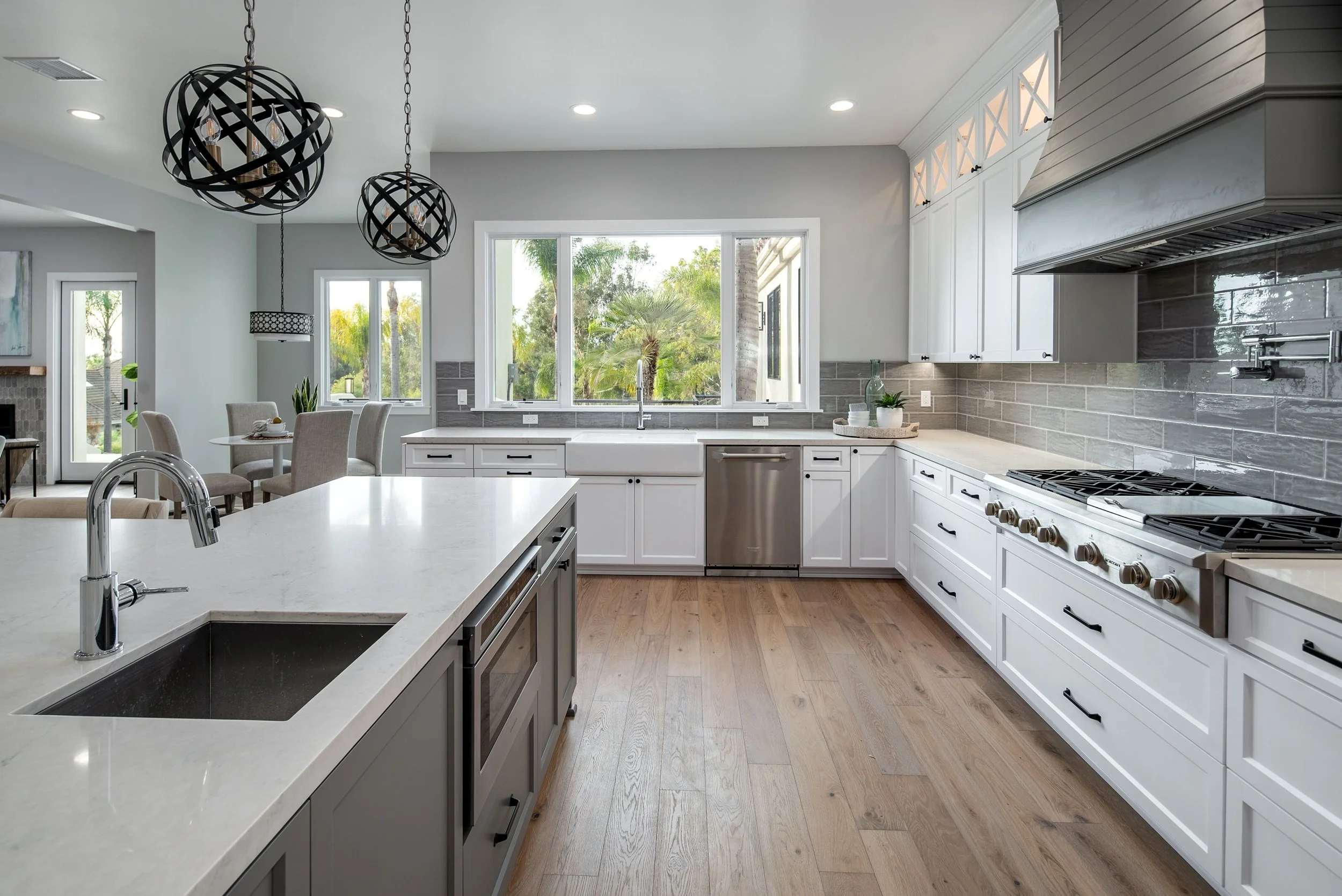 Modern kitchen with white cabinets, gray tile backsplash, large window, and wooden floors. A kitchen island with a sink in the foreground, and pendant lights hanging from the ceiling.