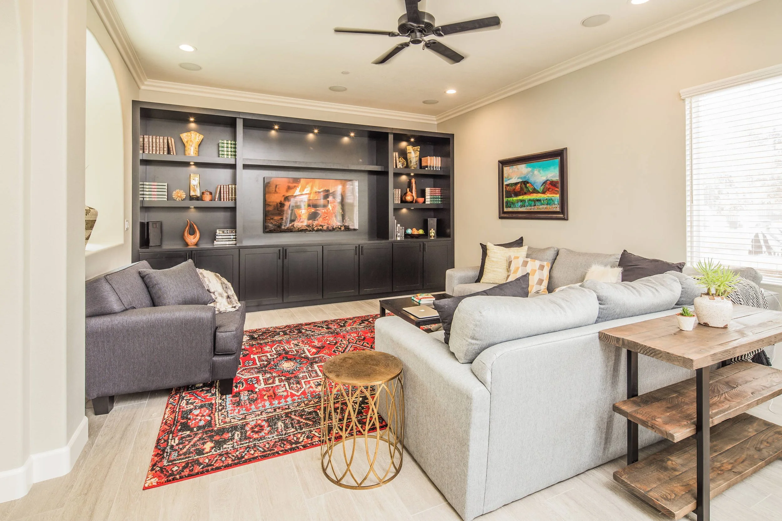 Living room with gray and beige sofas, a red patterned rug, a black built-in bookshelf with decorative items and fire place, wall art, a dining table, and a window with white blinds.