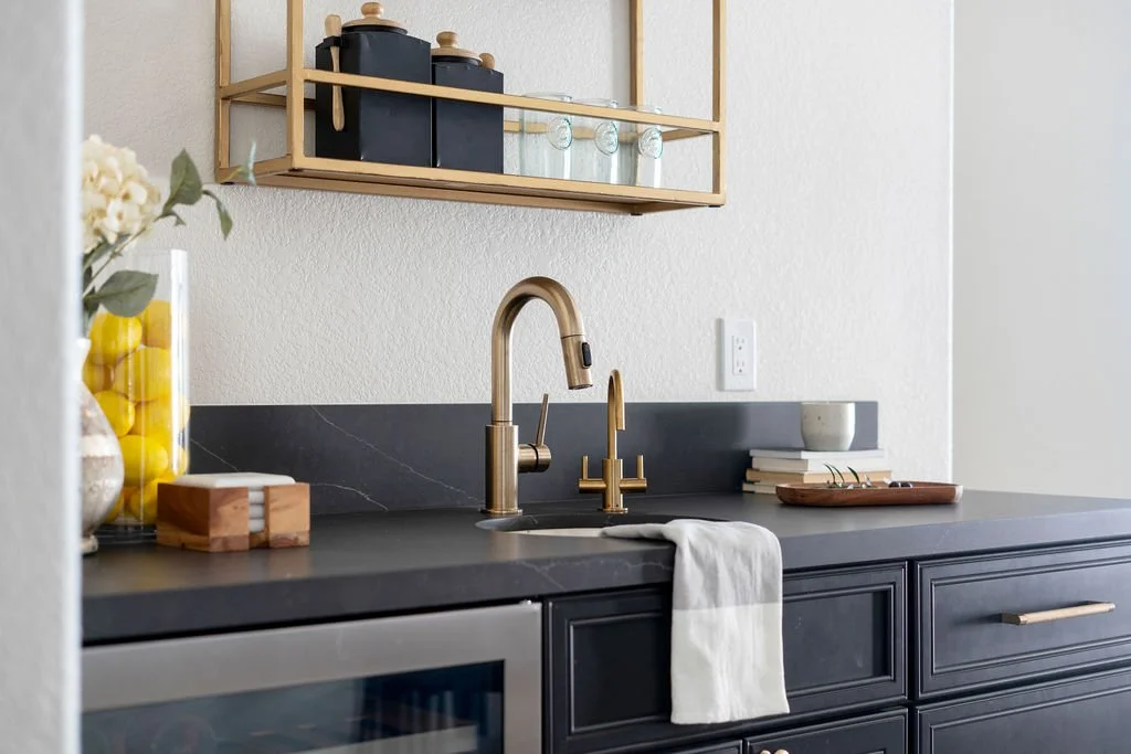 Modern kitchen sink with brass faucet, black countertop, and open wooden shelf holding jars and glassware.