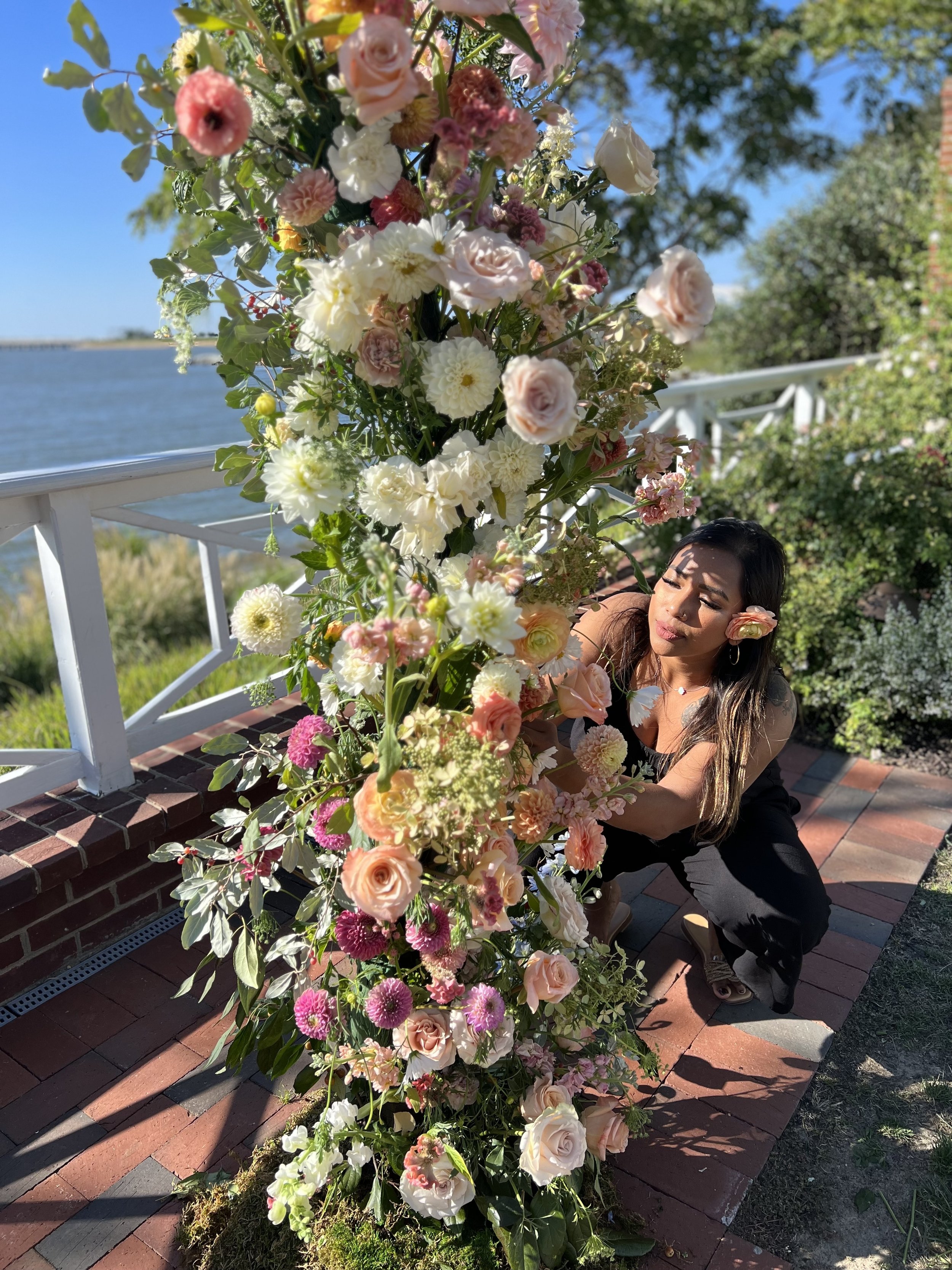 A woman in black clothing is arranging a large bouquet of various flowers, including roses, dahlias, and other blossoms, outdoors on a brick pathway near a body of water with trees in the background on a sunny day.
