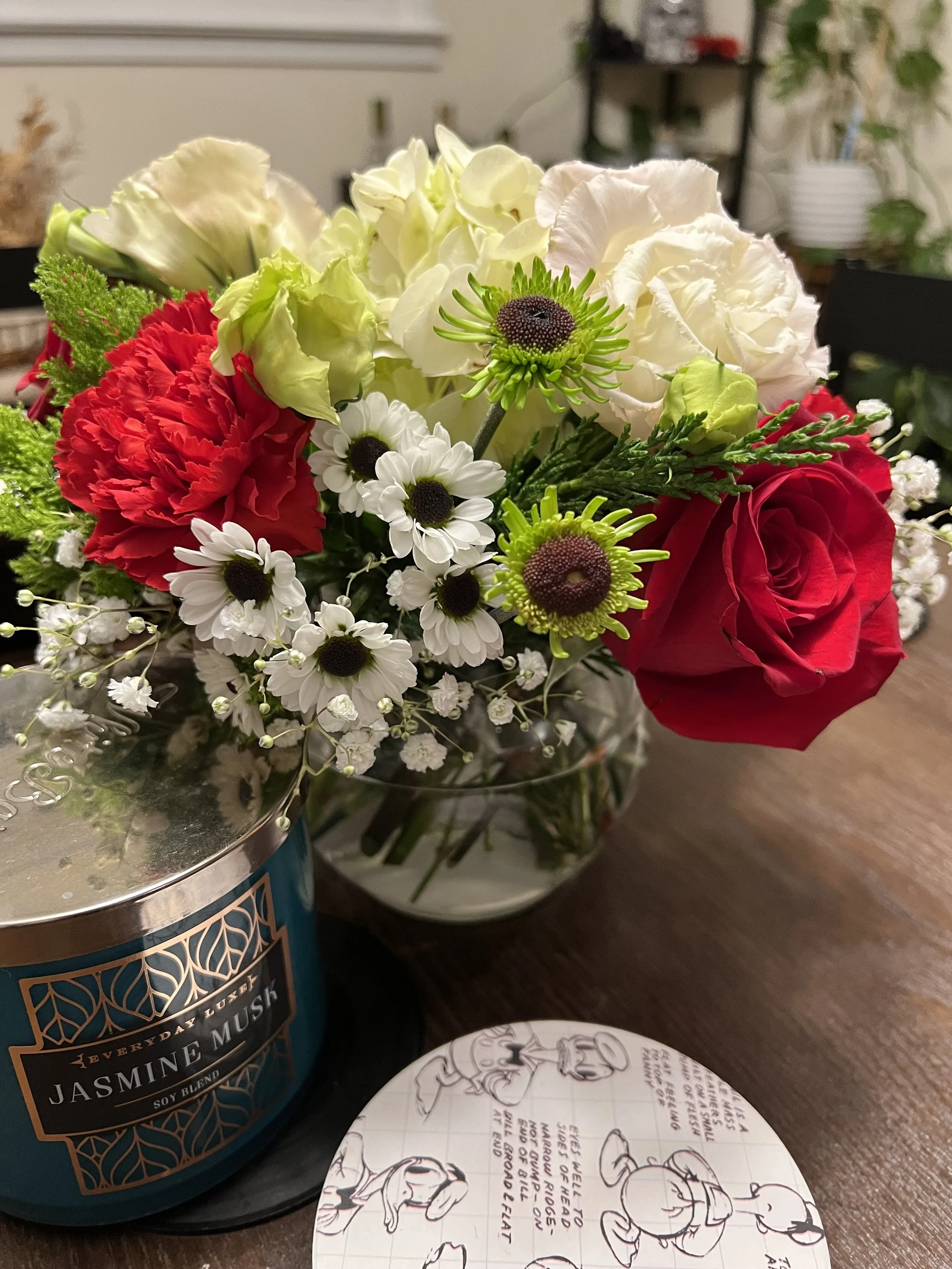 A colorful bouquet of flowers including red roses, white carnations, white daisies, and green accents in a glass vase, placed on a wooden table next to a decorative candle and a notebook with sketches.