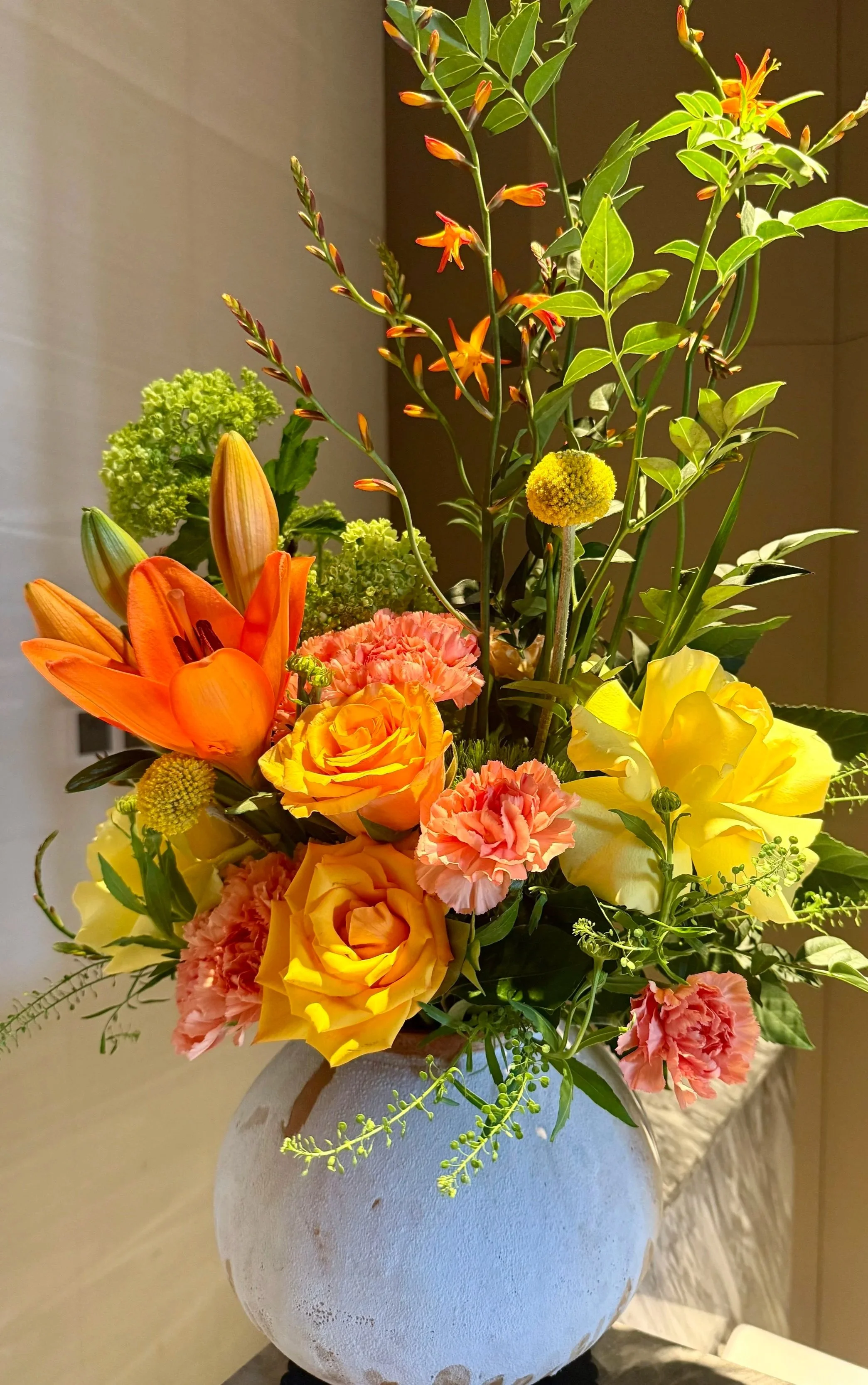 A colorful flower arrangement featuring yellow, orange, and pink blooms in a white textured vase.