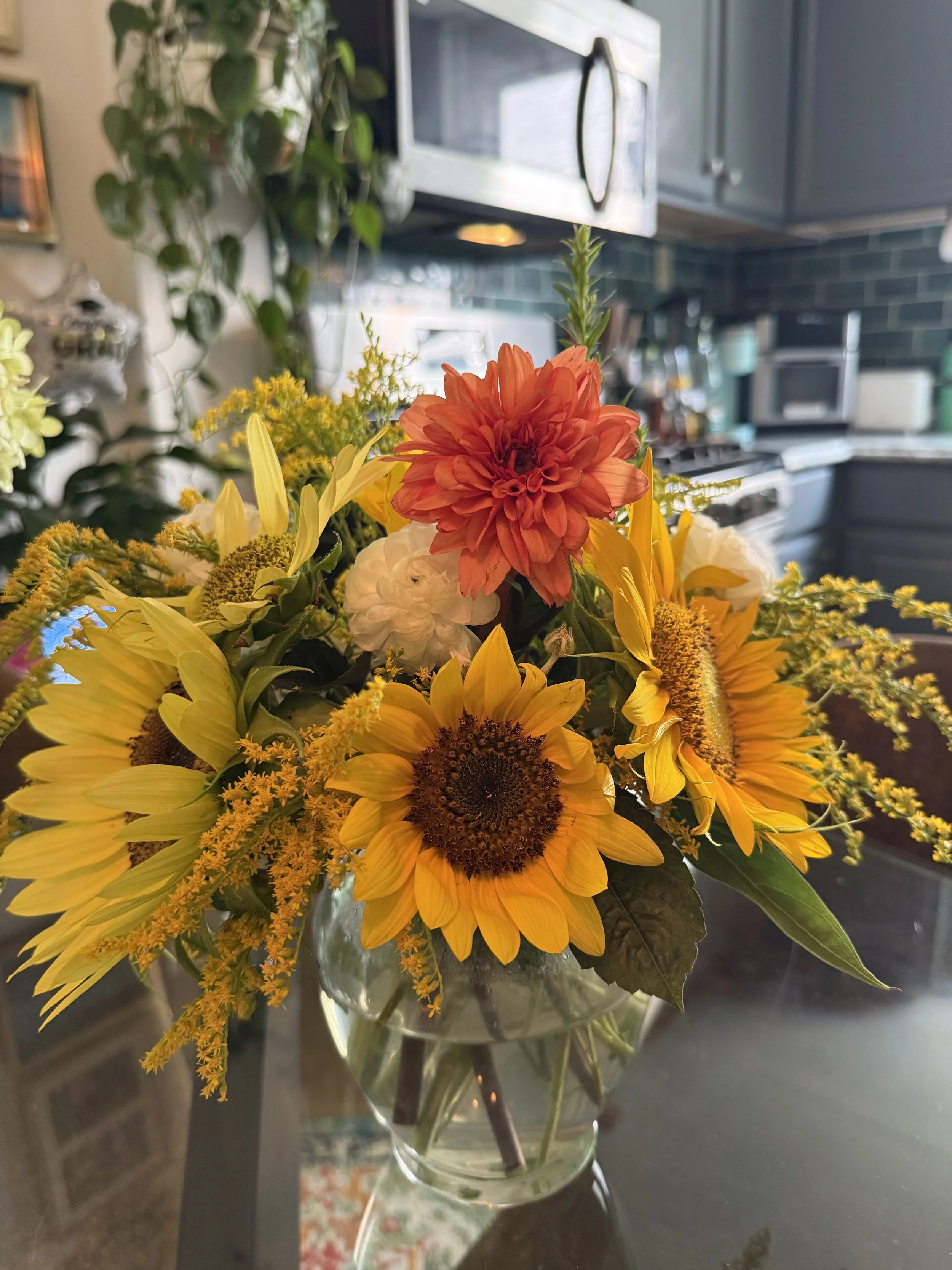 A colorful bouquet of flowers including sunflowers, a red/orange dahlia, white flowers, and yellow accents in a glass vase on a kitchen table.