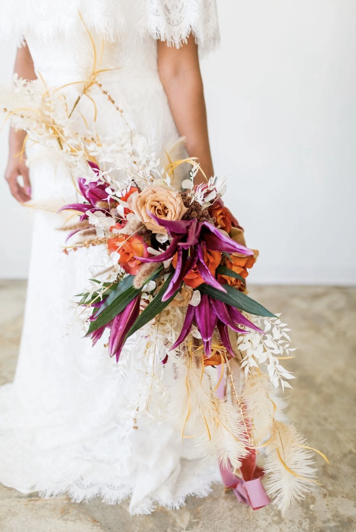 A woman in a white dress holding a large, vibrant bouquet of assorted flowers, including pink lilies, orange roses, beige roses, and dried white and beige foliage, with a pink ribbon at the bottom of the bouquet.