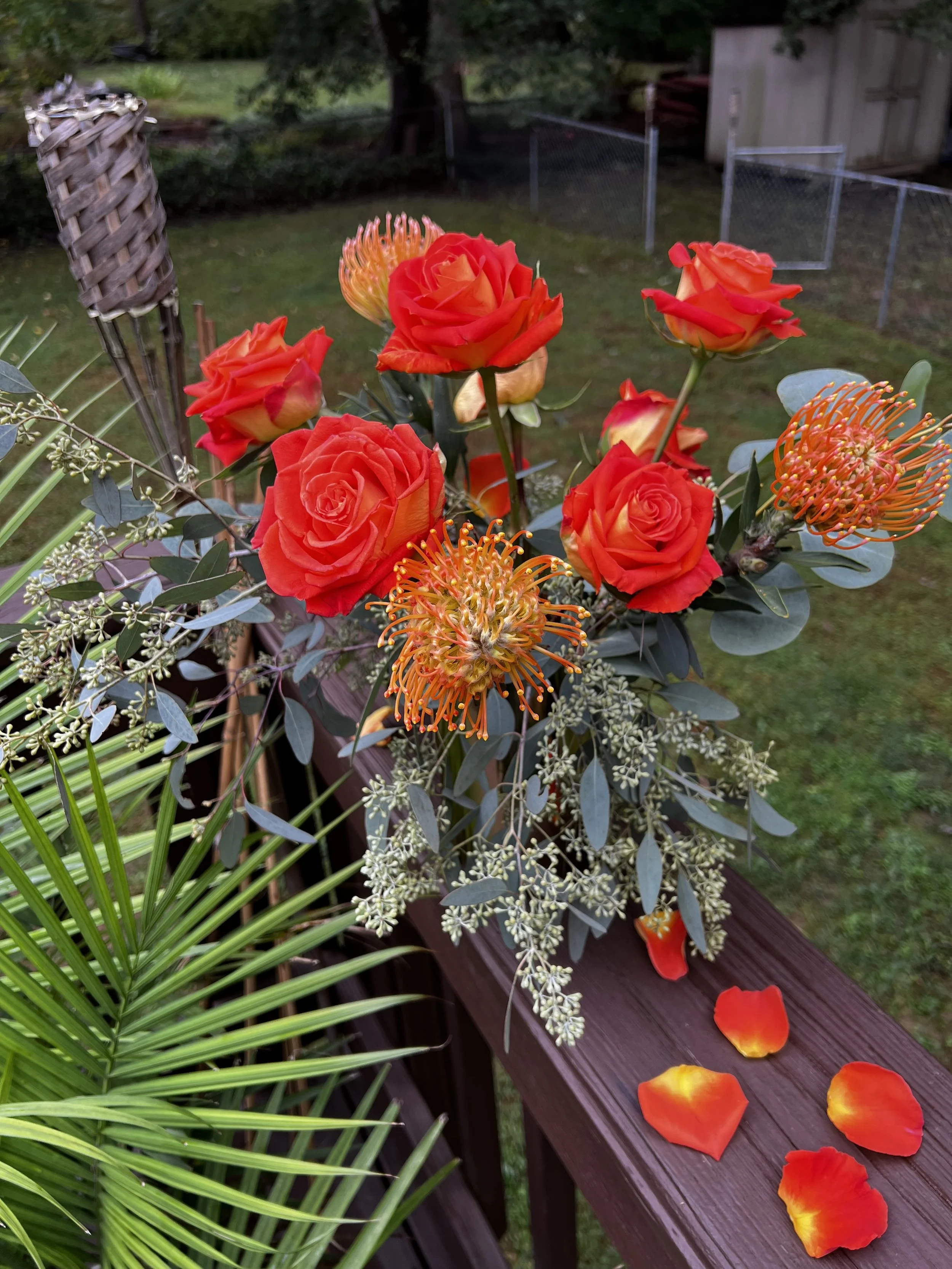 A flower arrangement with red roses, orange pincushion proteas, and greenery, placed on a wooden railing with fallen petals, outdoors in a backyard with a grassy yard and a fence.
