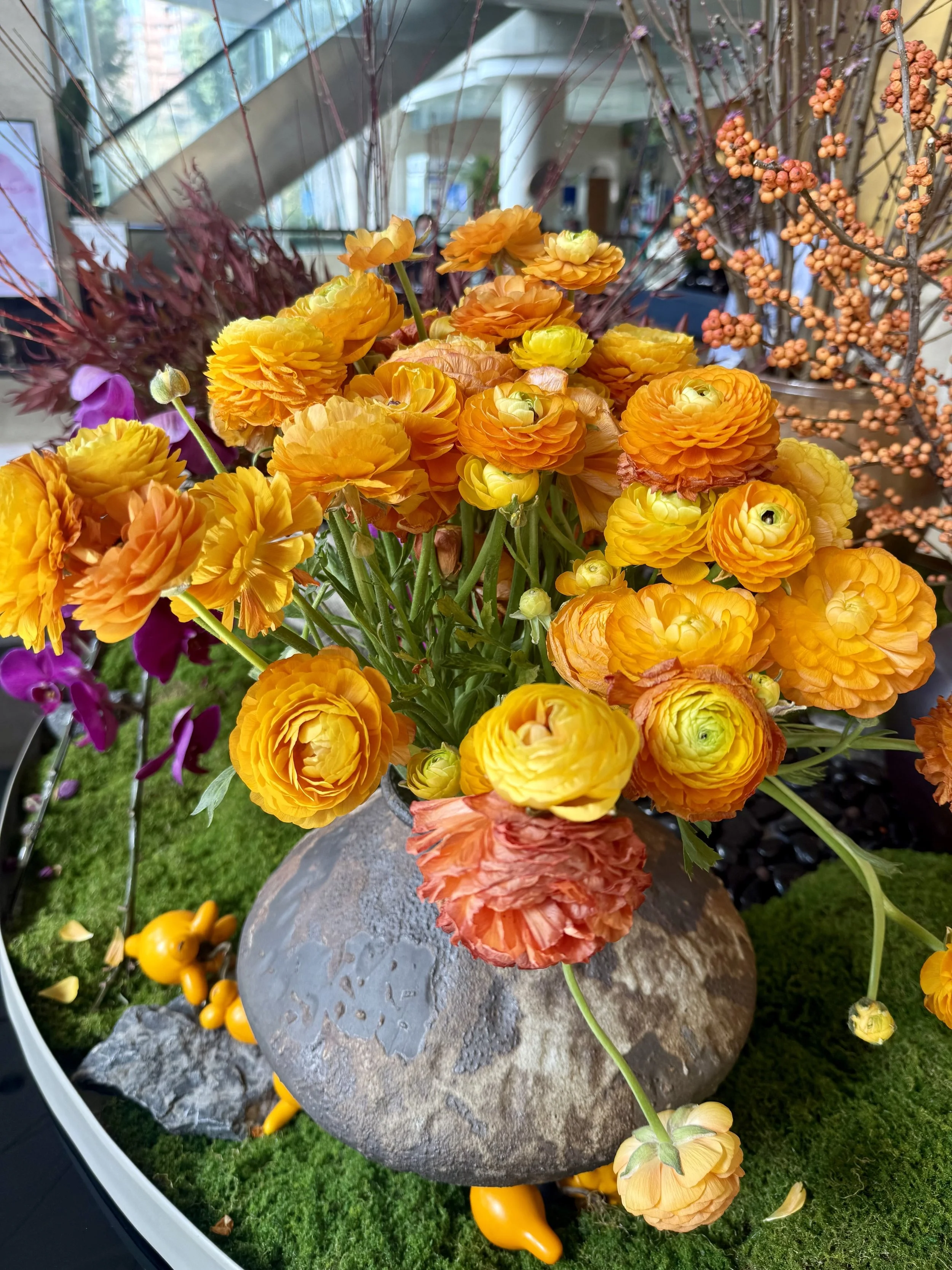 A vibrant bouquet of orange and yellow ranunculus flowers arranged in a rustic gray stone vase, with purple orchids, orange berries, and greenery in the background.