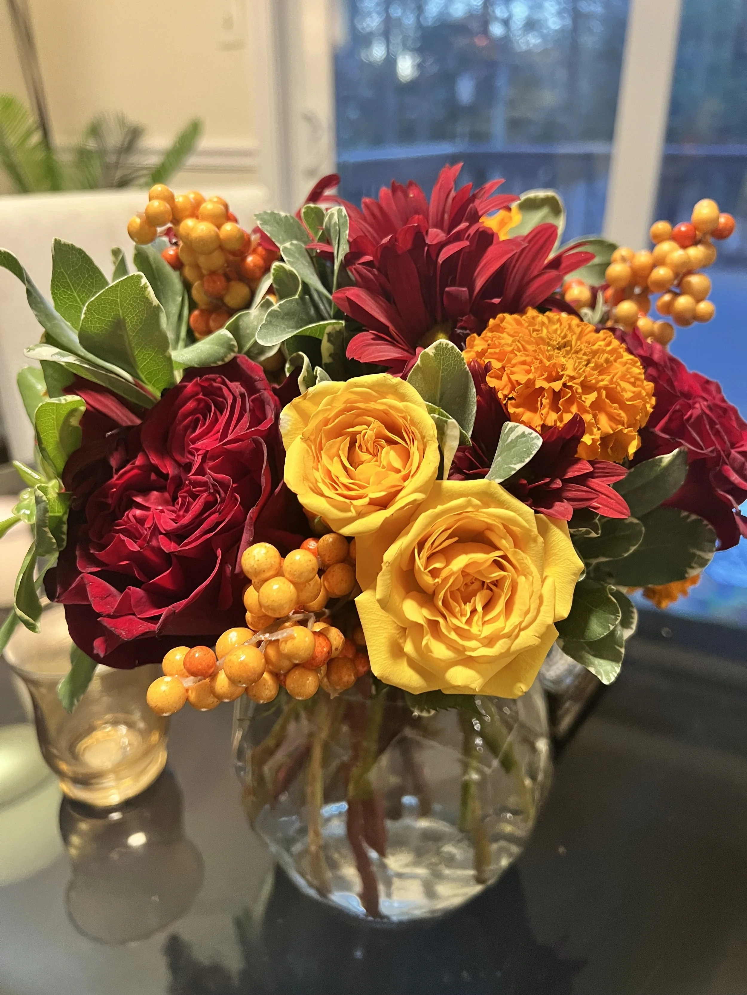 A colorful bouquet of flowers in a clear glass vase on a dark surface, featuring yellow roses, red roses, marigolds, red daisies, and orange berries, with a blurred background of a window and outdoor trees.