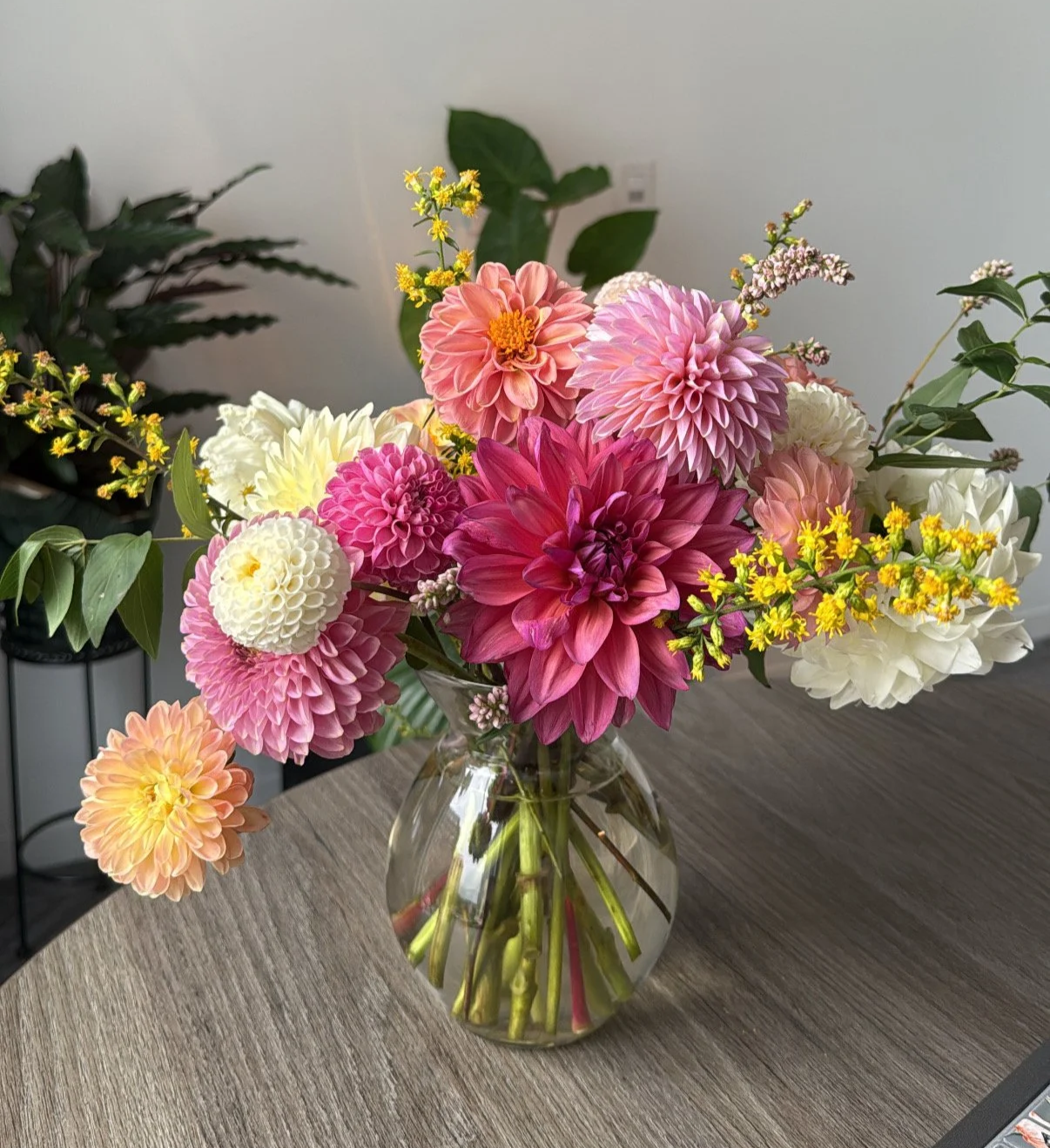 A glass vase filled with a colorful bouquet of various flowers including dahlias in shades of pink, white, and peach, along with yellow accent flowers, placed on a wooden table.